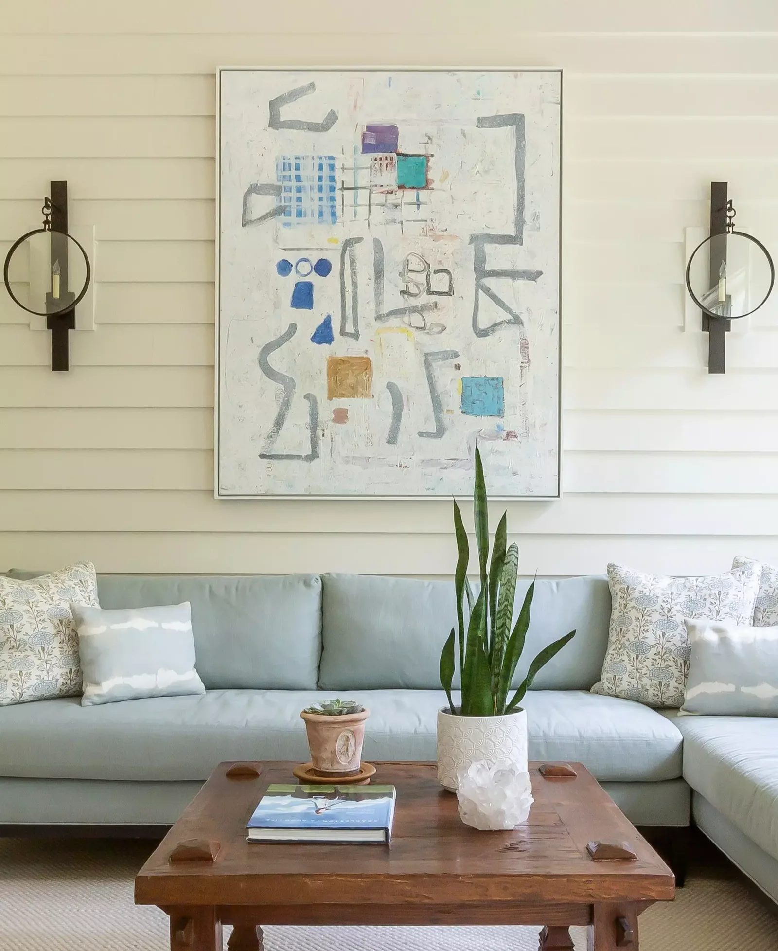 Living room with a light gray sofa, abstract artwork, and two black wall sconces, with a wooden coffee table and a tall plant.