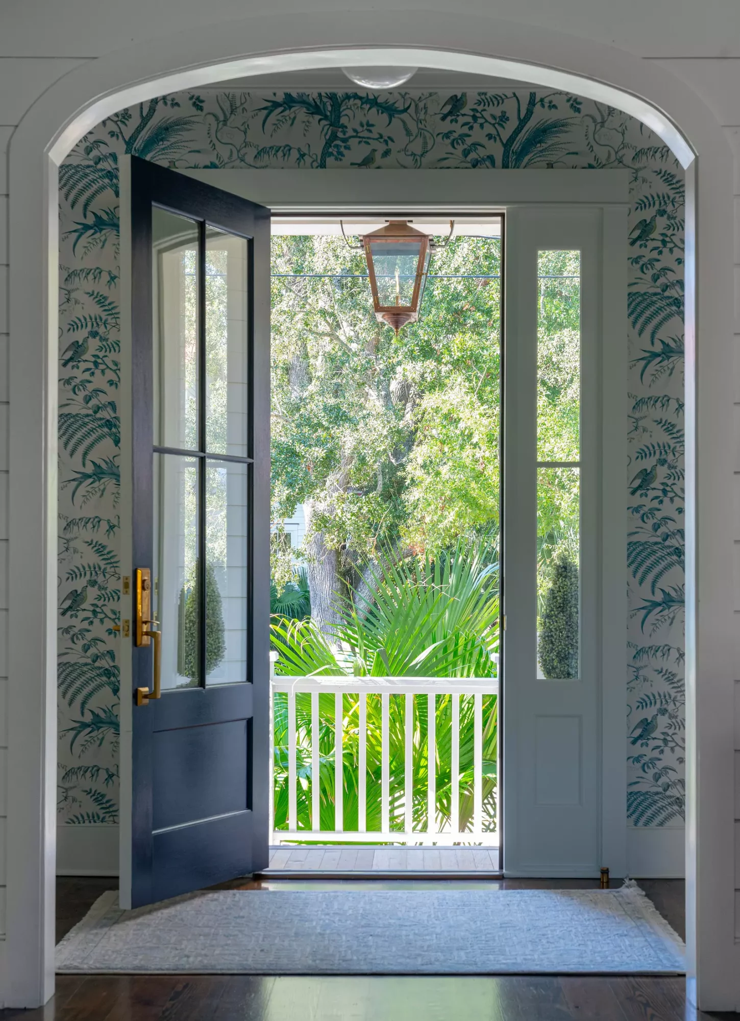Open door leading to a balcony with greenery and trees outside, framed by patterned wallpaper and an arched doorway.