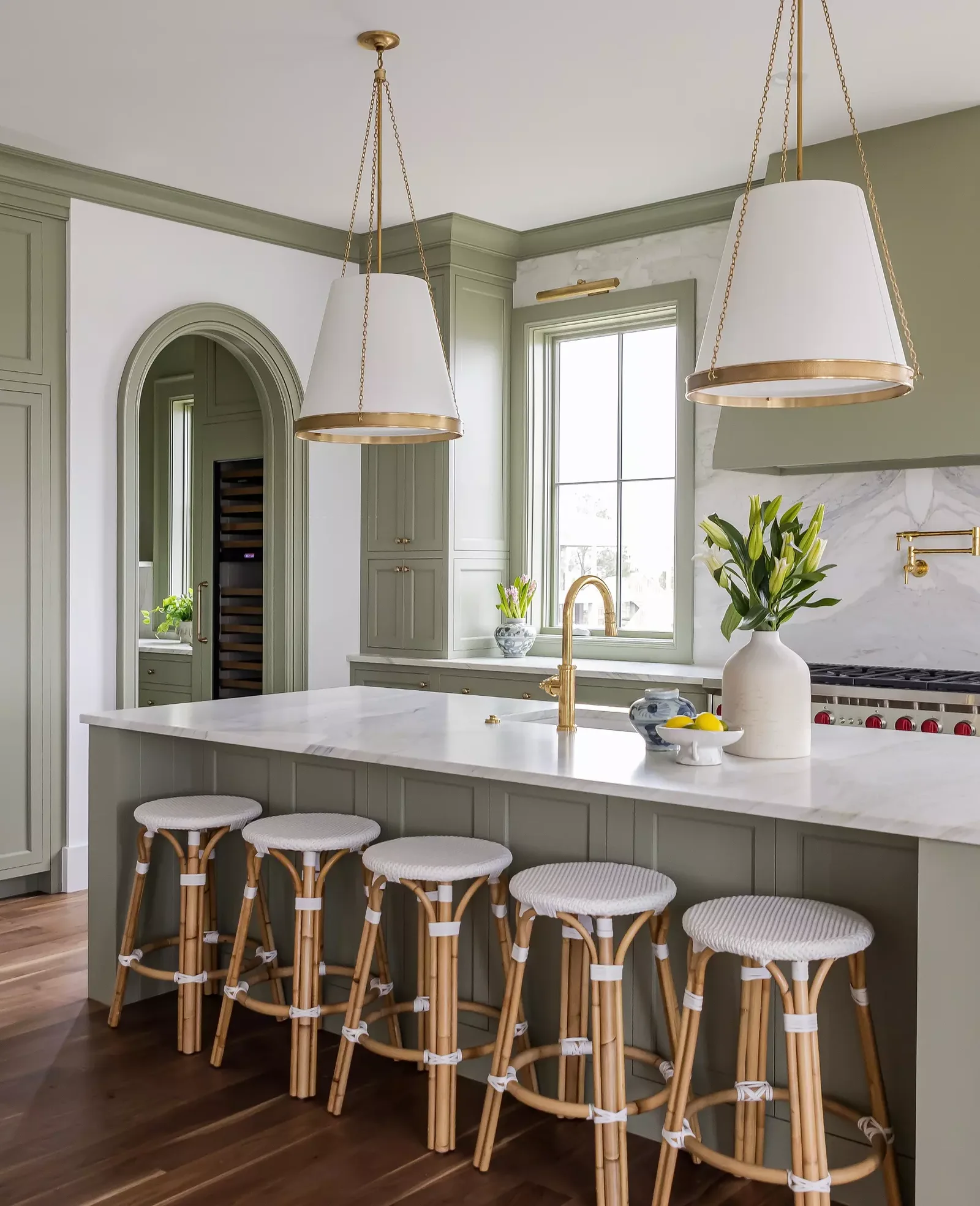 Kitchen with green cabinets, white marble island, gold fixtures, and hanging pendant lights, with a window and plants in the background.
