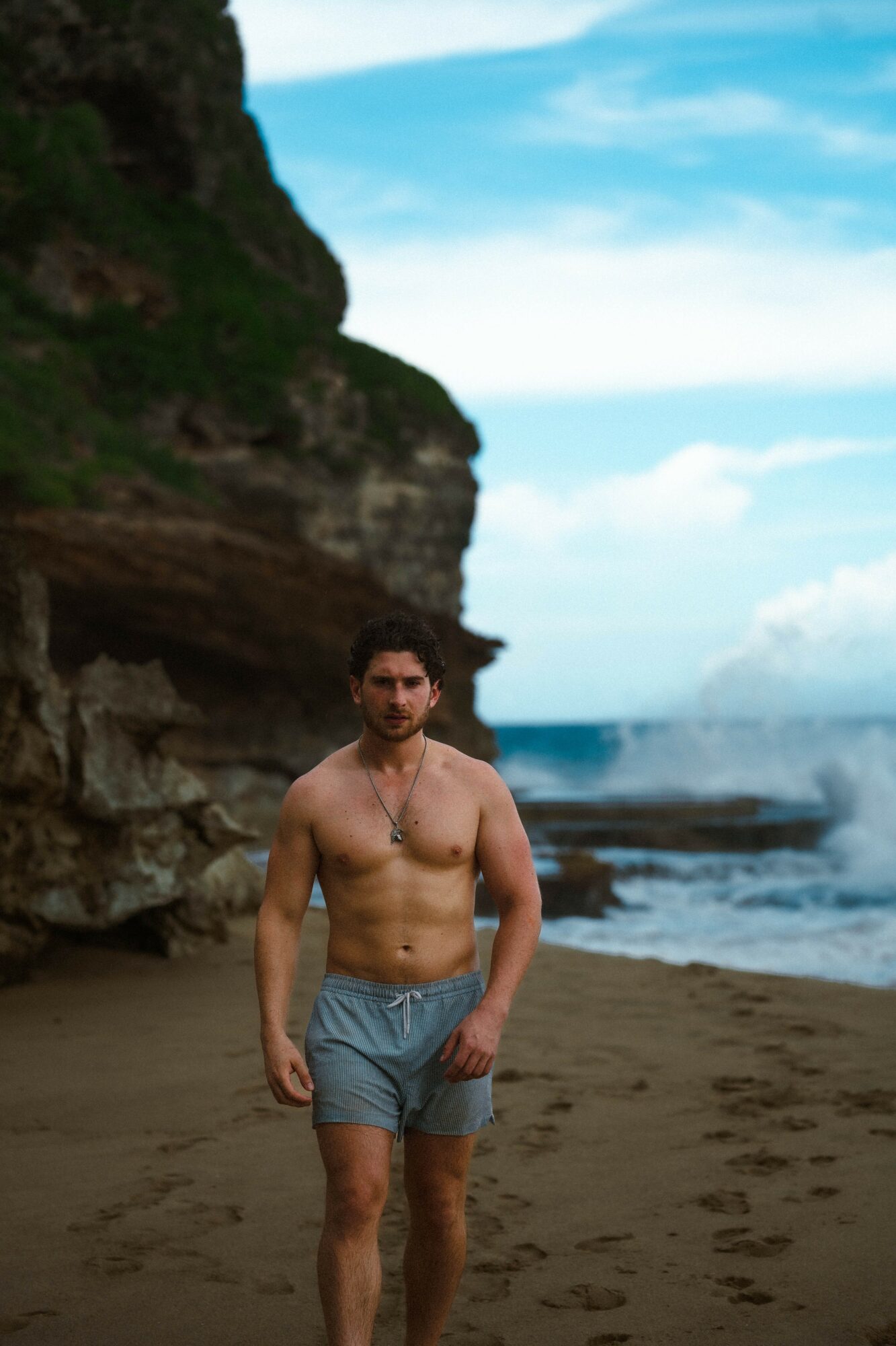 A shirtless man in shorts walking on a beach with cliffs and ocean in the background.