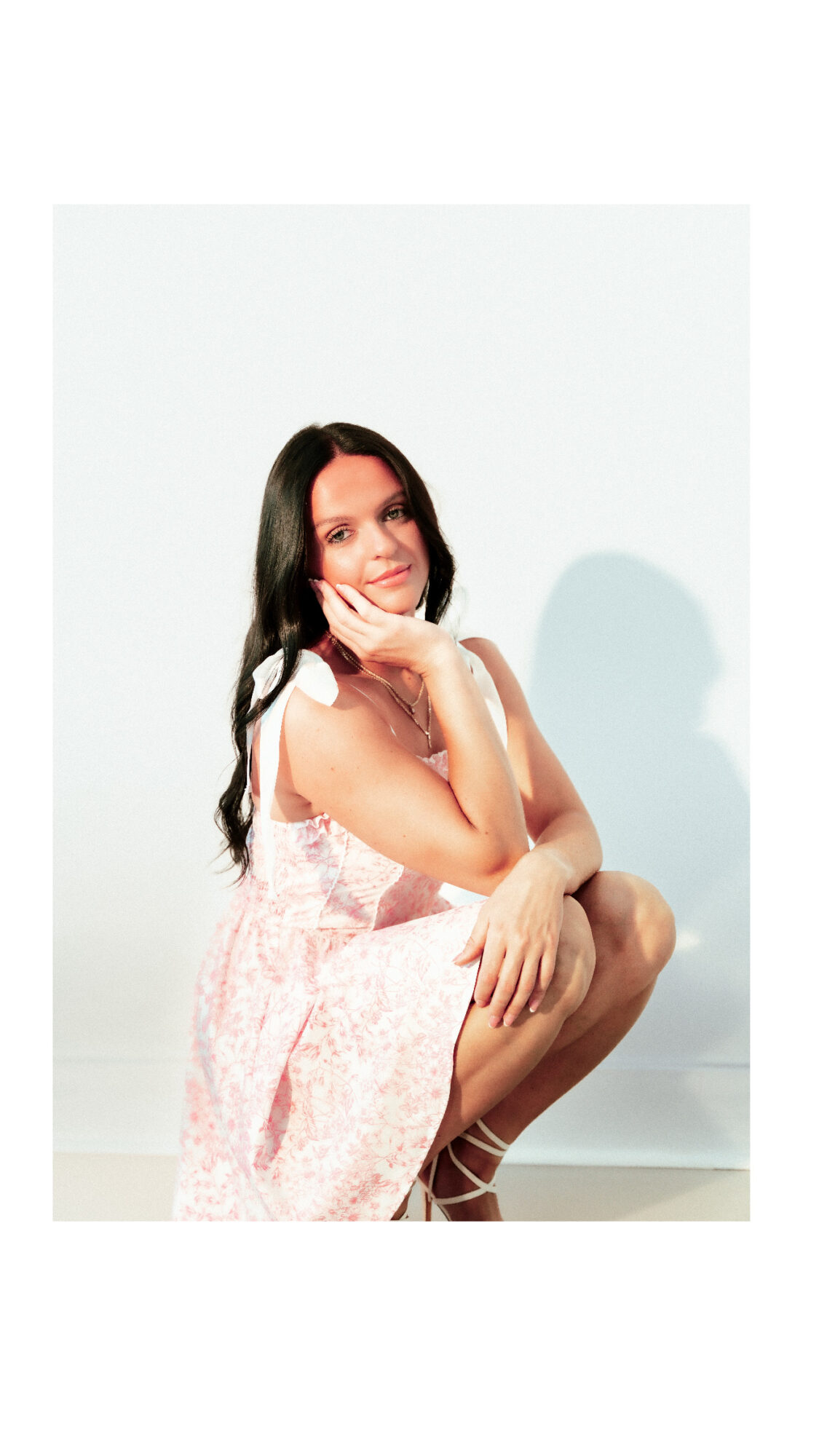 Woman with long dark hair crouching, wearing a pink dress, smiling, with a light background and shadow, hand near face.