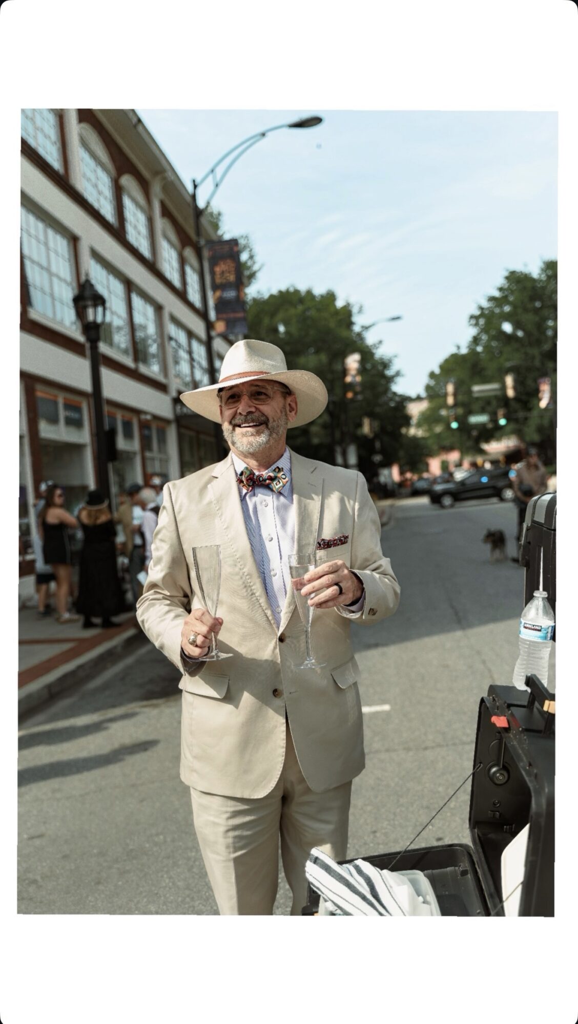 Man in light suit and hat holding a drink on a city street, smiling, with buildings and trees in background.