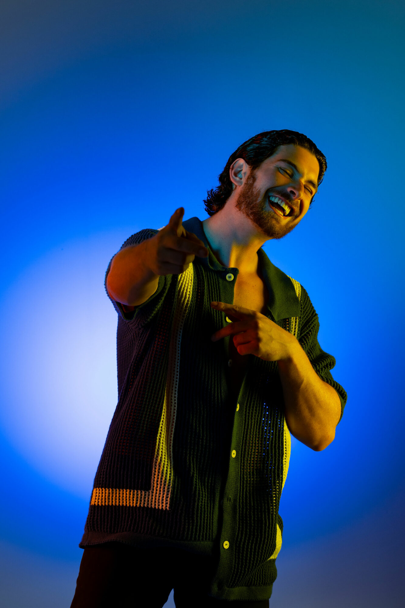 Young man with a beard smiling and pointing at the camera, standing against a blue background.