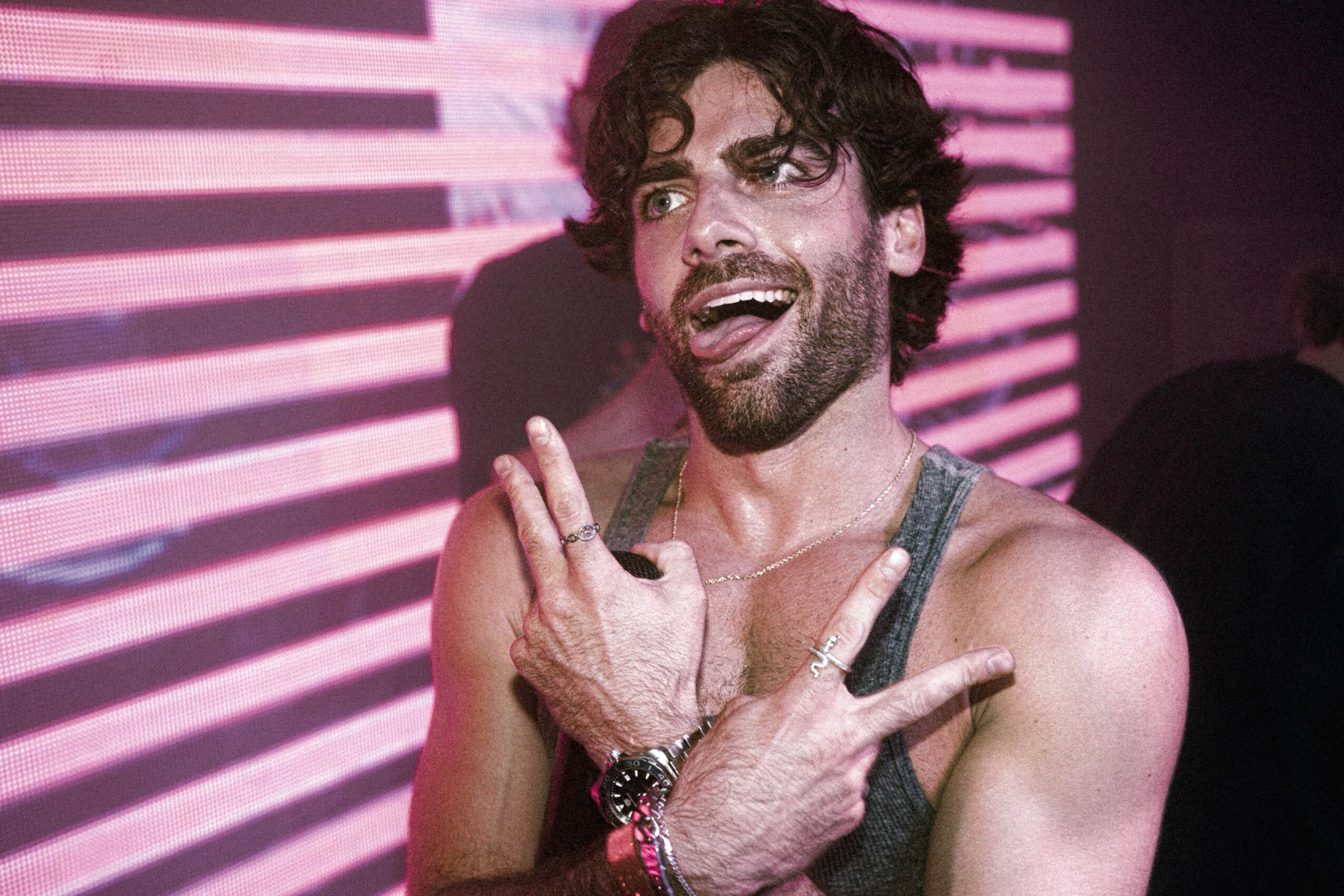 Man with dark curly hair and beard smiling, making peace signs with both hands, in front of pink and purple striped background.