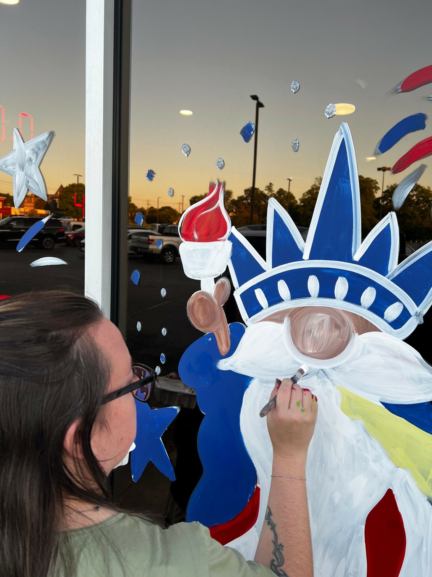 Person dressed as the Statue of Liberty with a crown and torch, standing outdoors during sunset, with a woman in the foreground.