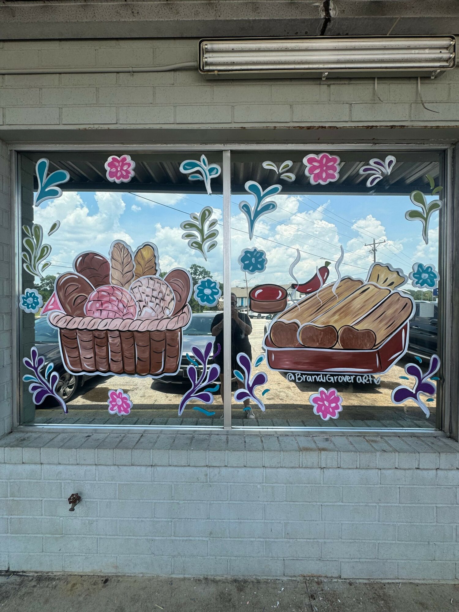 Decorative window with painted cakes, flowers, and clouds, viewed from outside, with a blue sky background.
