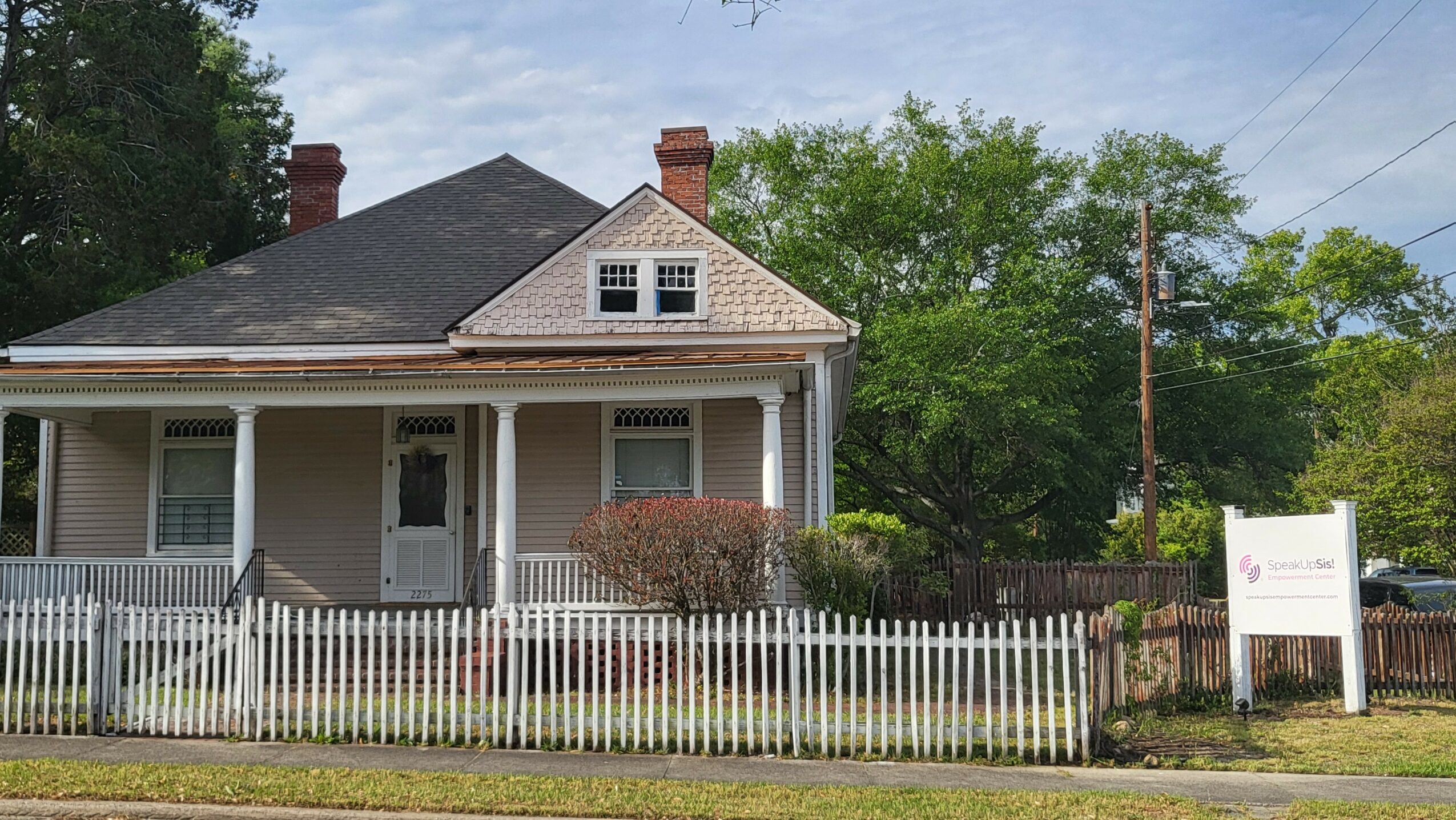 A two-story house with a porch, white picket fence, and a sign on the lawn, surrounded by trees and greenery.