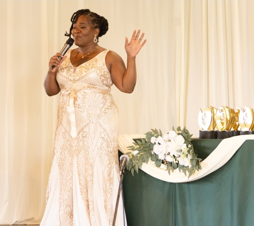 Woman in a white dress speaking into a microphone with her hand raised, standing near a table with flowers and awards.