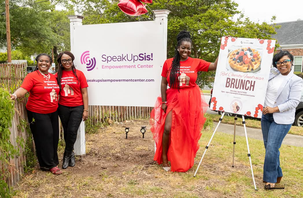 Four women standing outdoors near a sign and a brunch poster, two women in red dresses and two in casual attire, smiling.