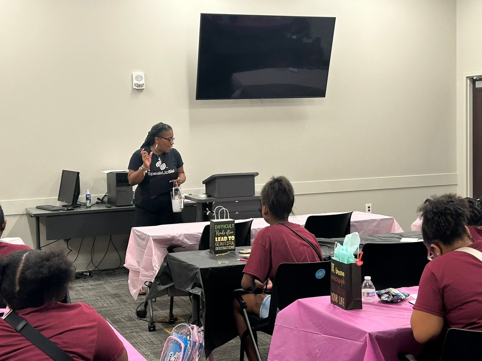 A woman stands at a table giving a presentation to seated people in a room with a large TV screen on the wall.