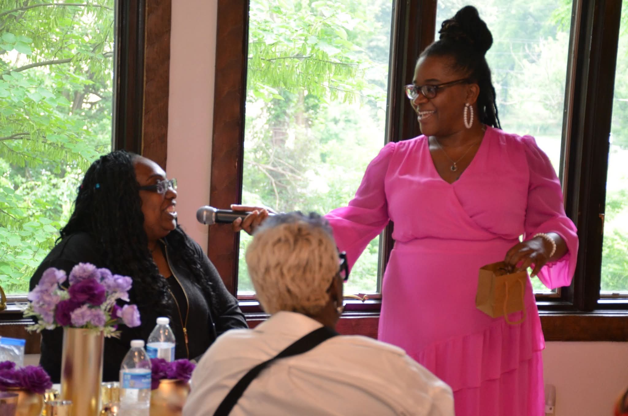 Woman in pink dress speaking into microphone at a table with three people, flowers, and drinks, with windows and greenery outside.