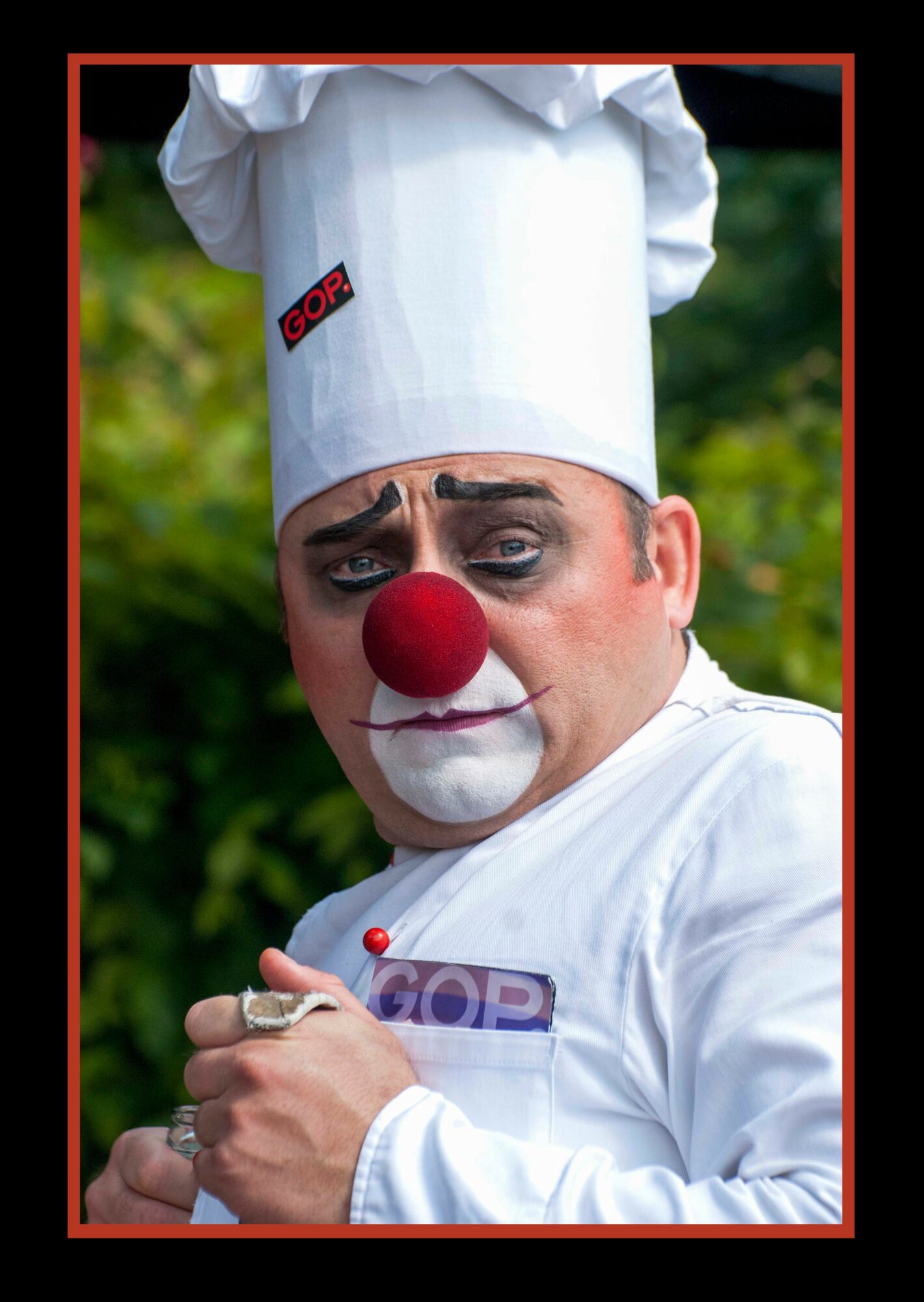 Person dressed as a clown with a red nose, white chef's hat, and white outfit, outdoors with green foliage background.