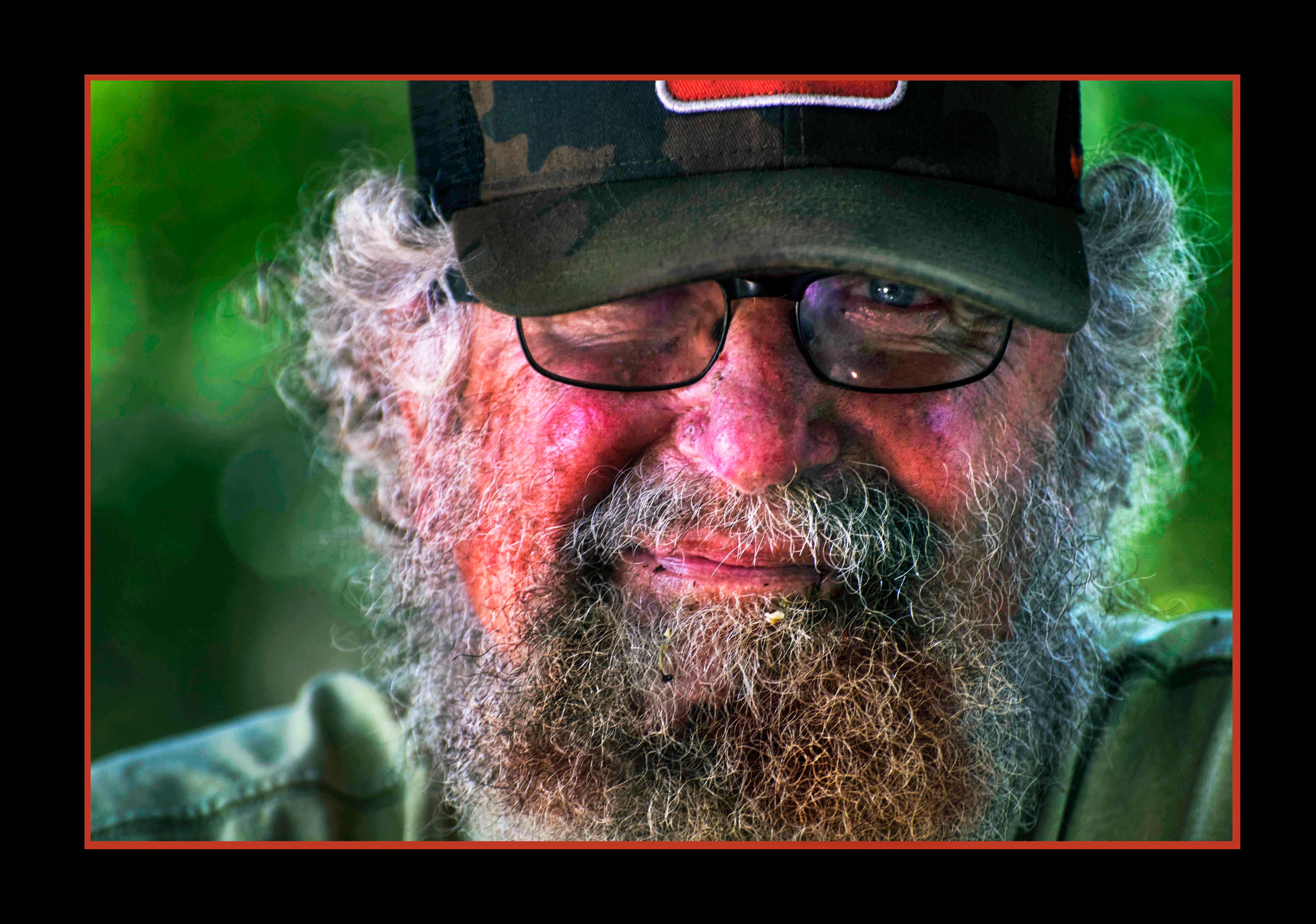 Close-up of an older man with glasses, a beard, and a cap, smiling outdoors with a blurred green background.