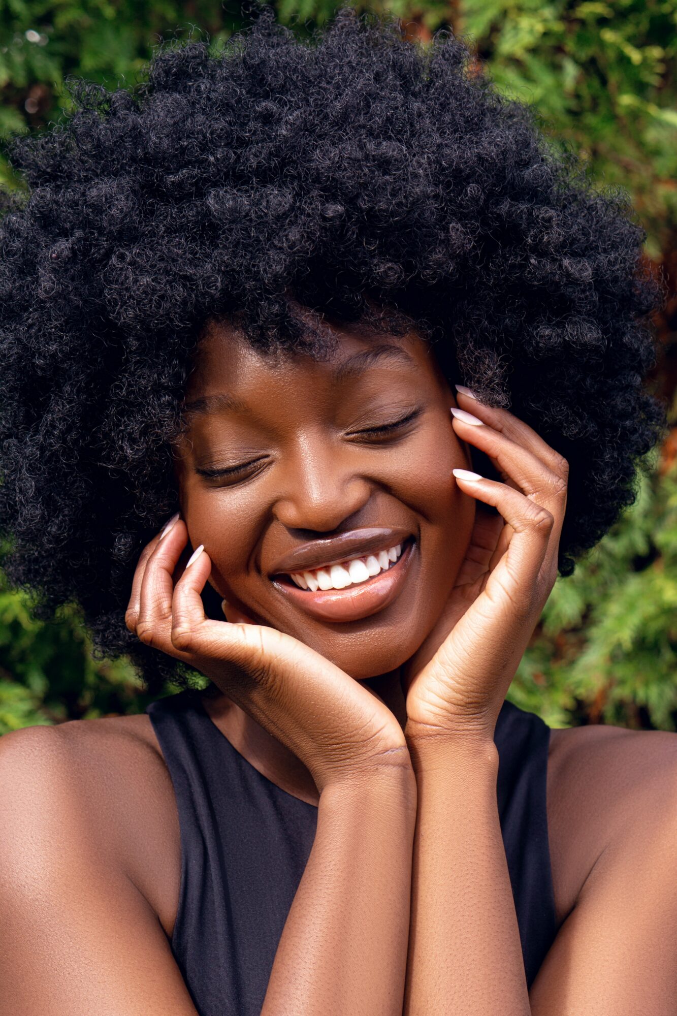 Smiling woman with natural curly hair outdoors, hands near face, wearing a black top.