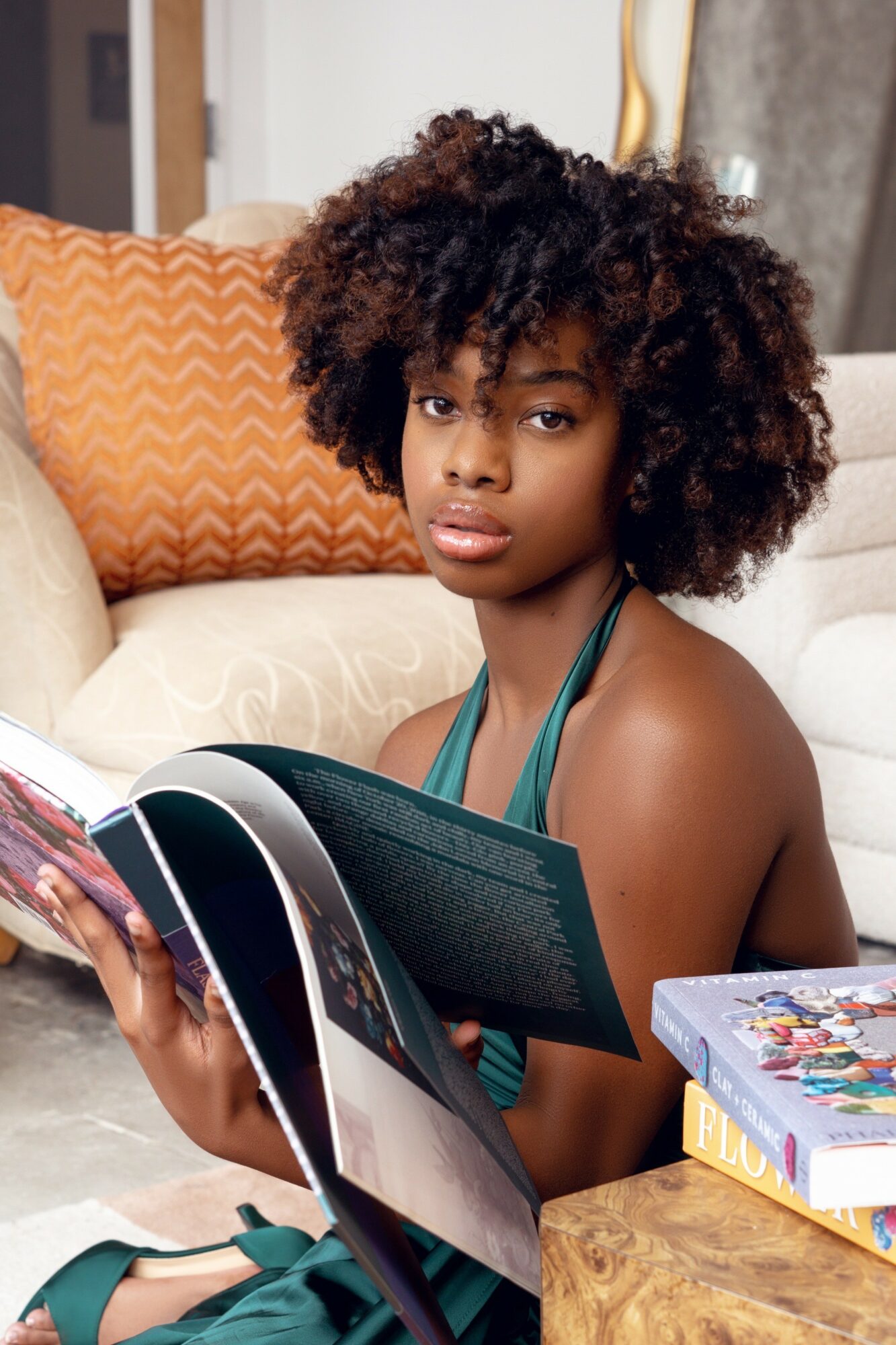 Young woman with curly hair sitting on a couch, holding a magazine, with books nearby, in a cozy living room.