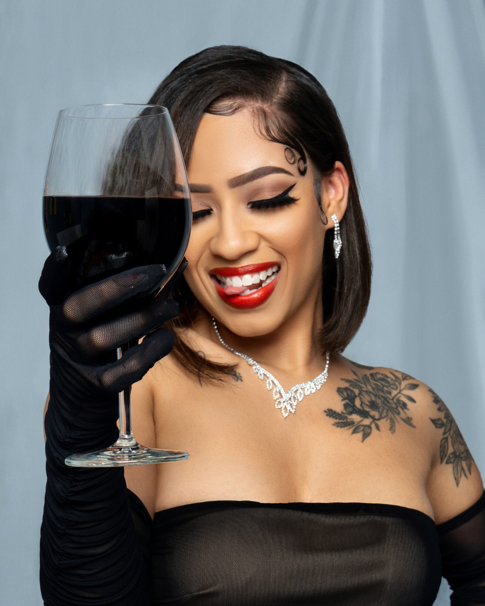Woman with dark hair, makeup, and tattoos, smiling and holding a glass of red wine, wearing jewelry and a black strapless top.