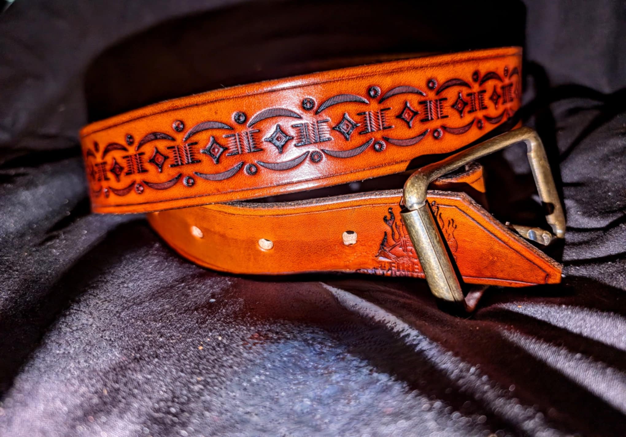 Orange belt with black decorative pattern and a gold buckle, resting on dark fabric.
