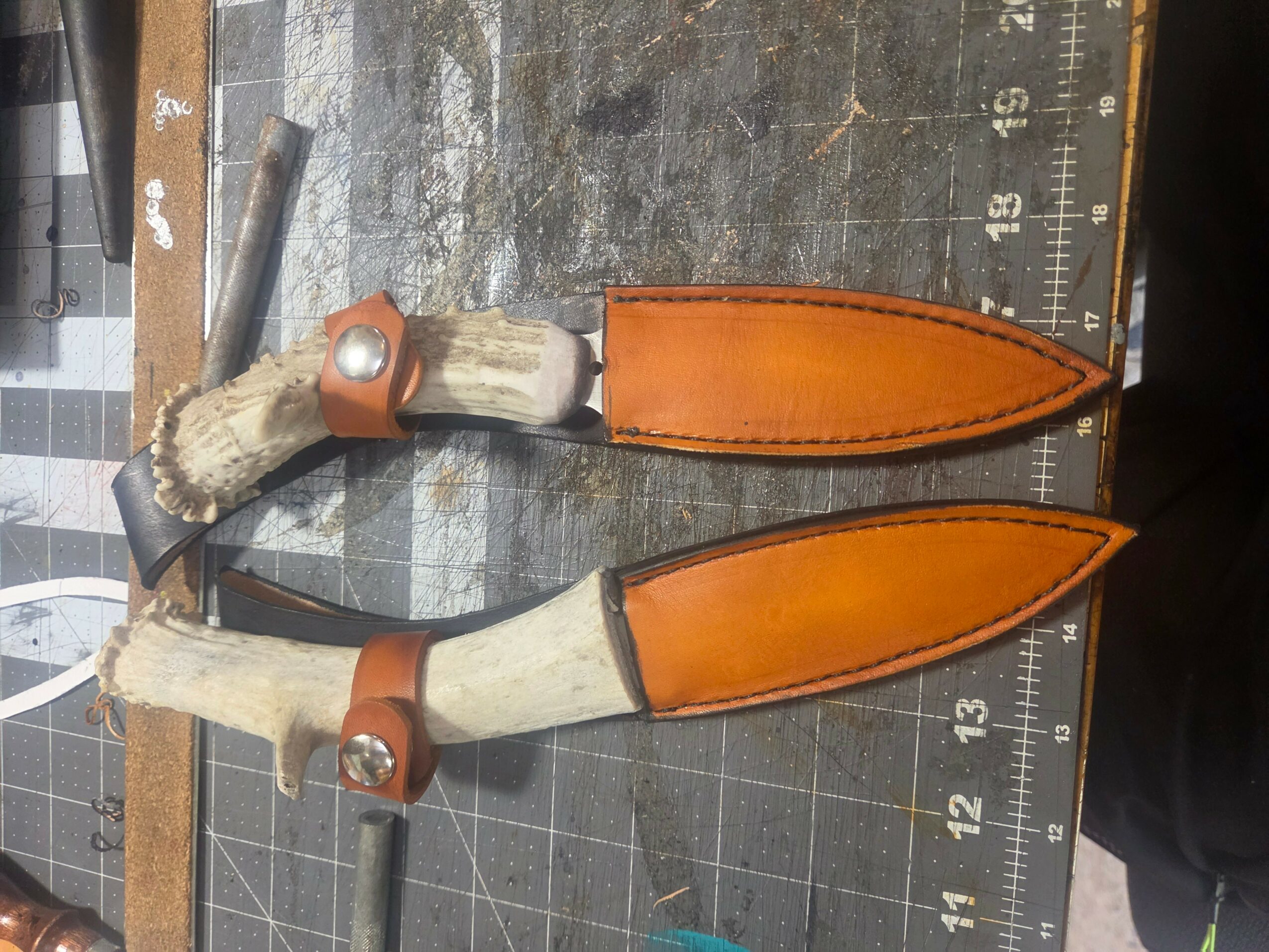 Two knives with wooden handles and orange leather sheaths, placed on a cutting mat with measurement markings.
