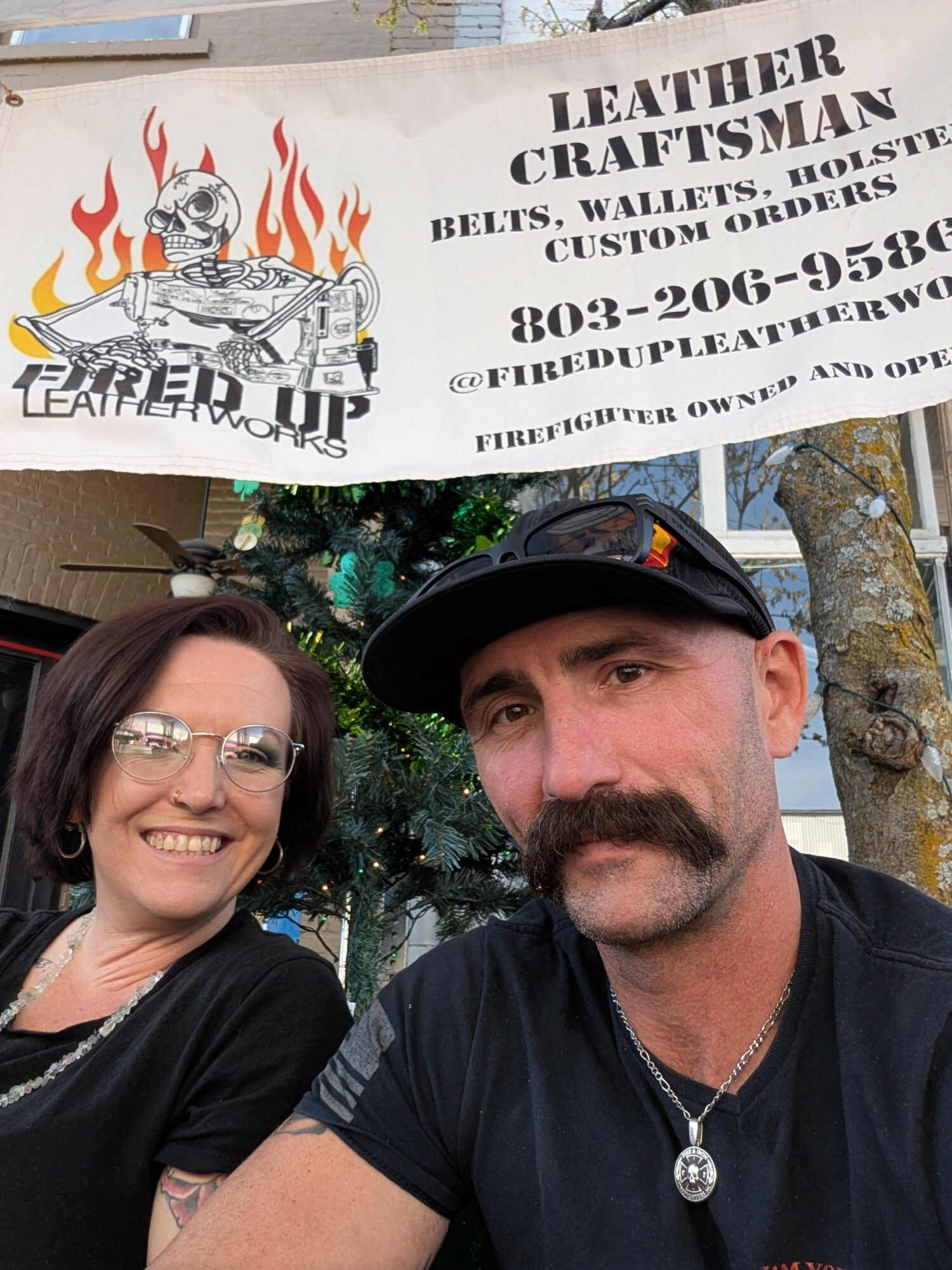 Two people smiling outdoors in front of a sign for leather crafts, with trees and a Christmas tree in the background.