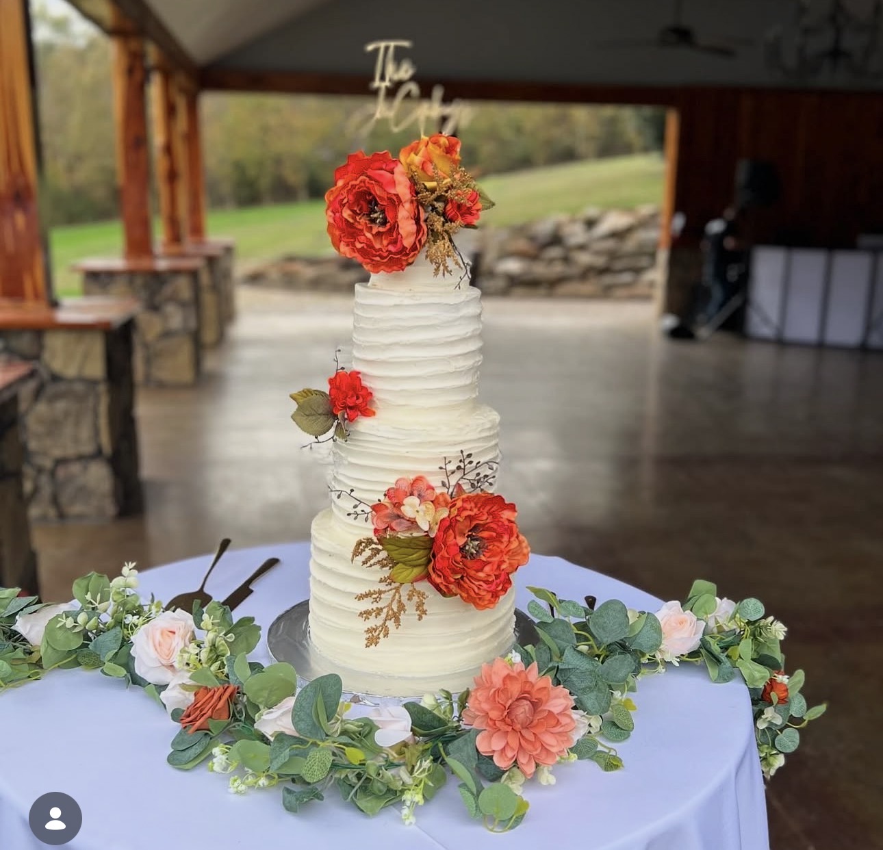 Three-tiered white wedding cake decorated with red and pink flowers, surrounded by greenery on a table.