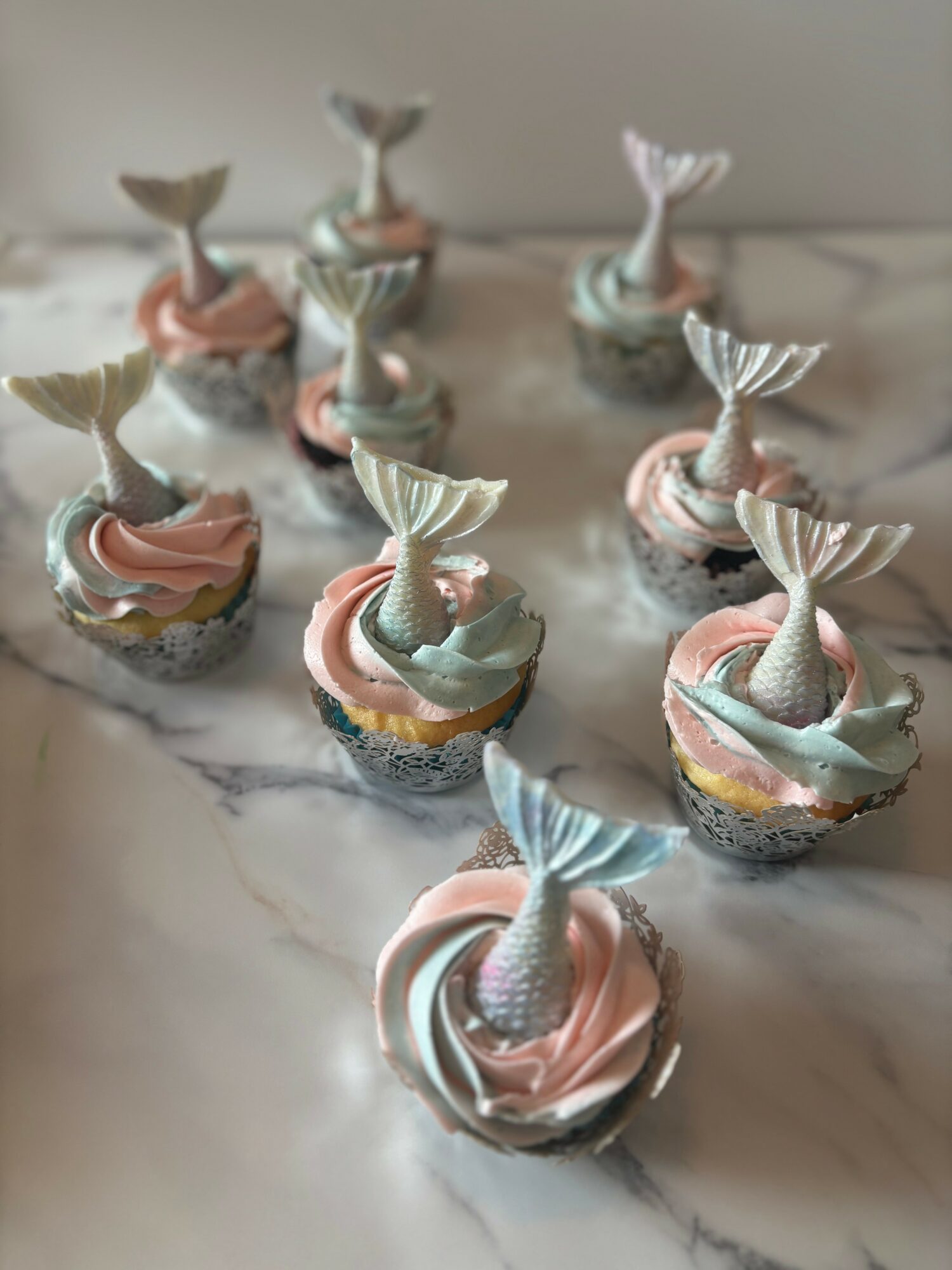 Nine cupcakes decorated with mermaid tail toppers and pastel-colored frosting on a marble surface.