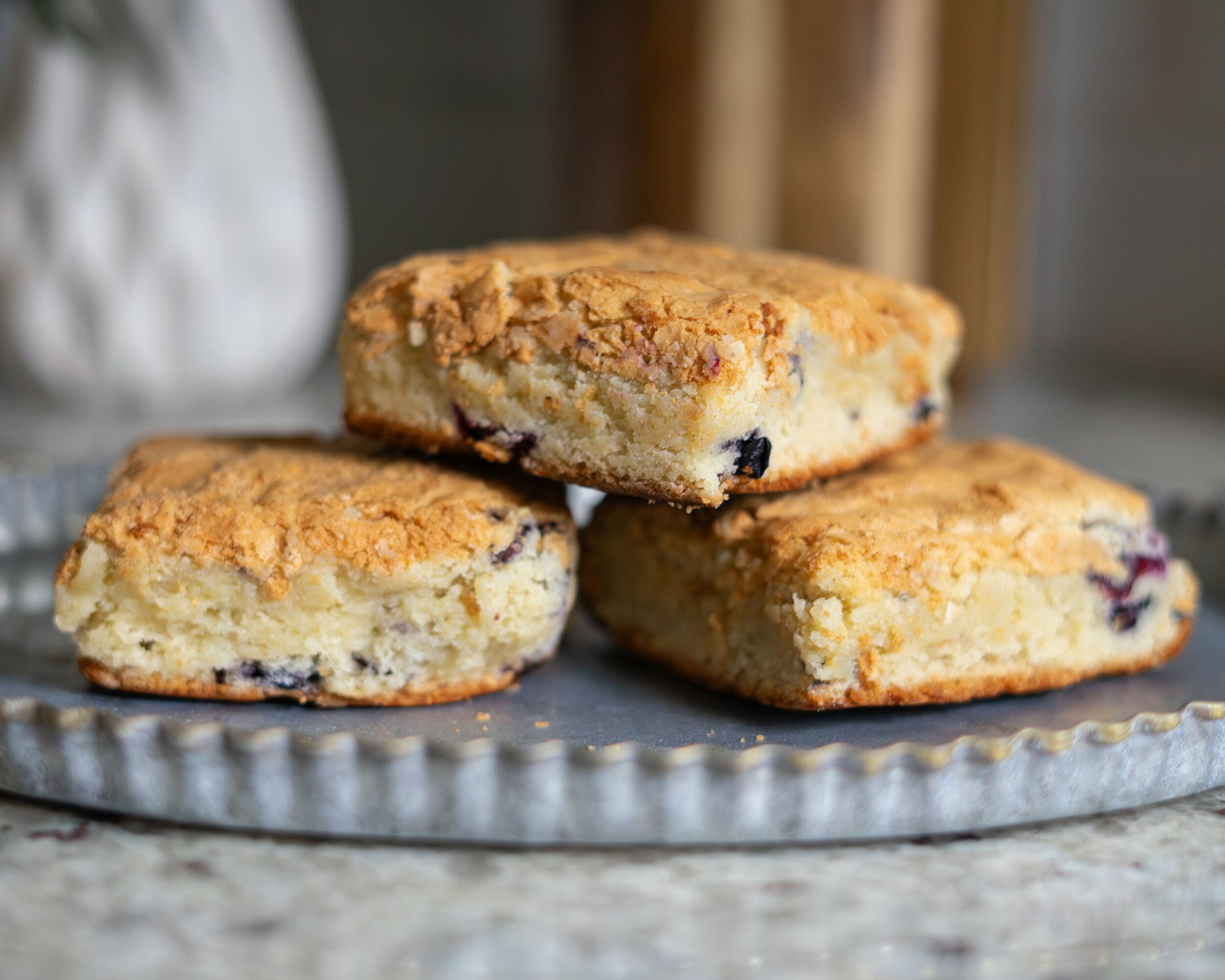 Three blueberry scones stacked on a silver tray, with a blurred background, showing golden-brown crust and blueberries inside.
