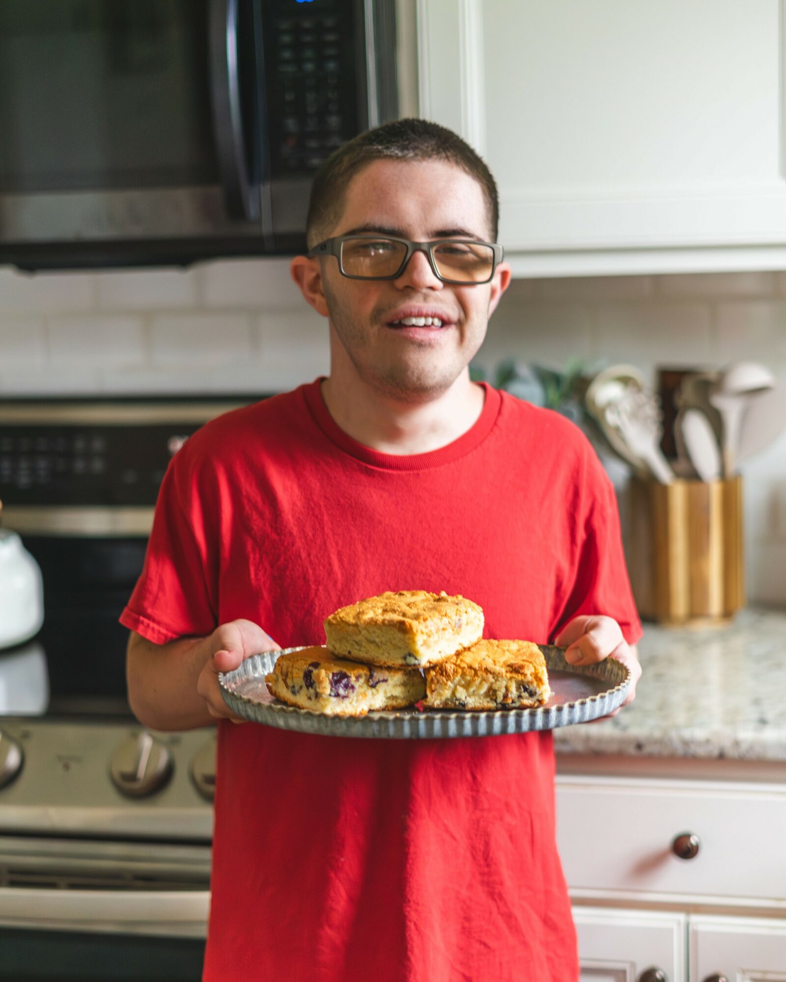 Young man with glasses in a red shirt holding a plate of baked goods in a kitchen.