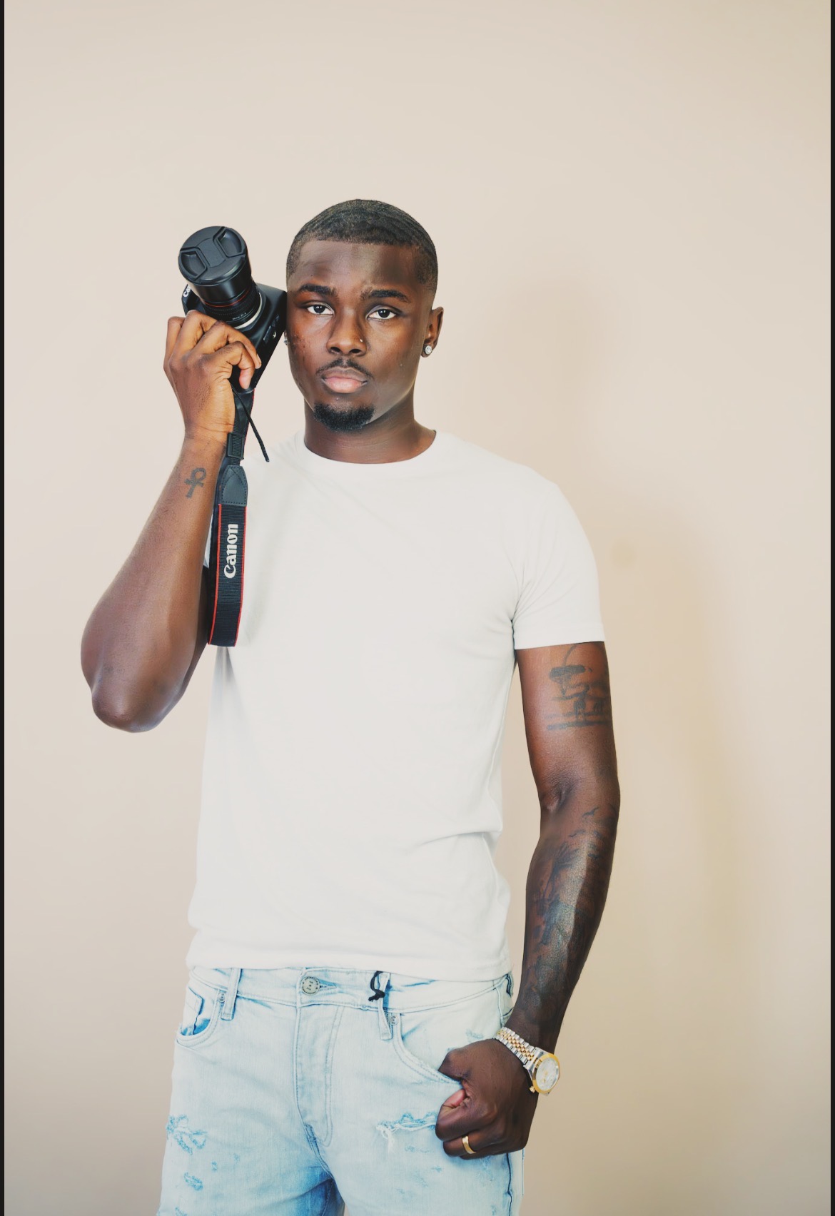 Young man with short hair holding a camera to his ear, wearing a white t-shirt and light blue jeans, standing against a plain background.