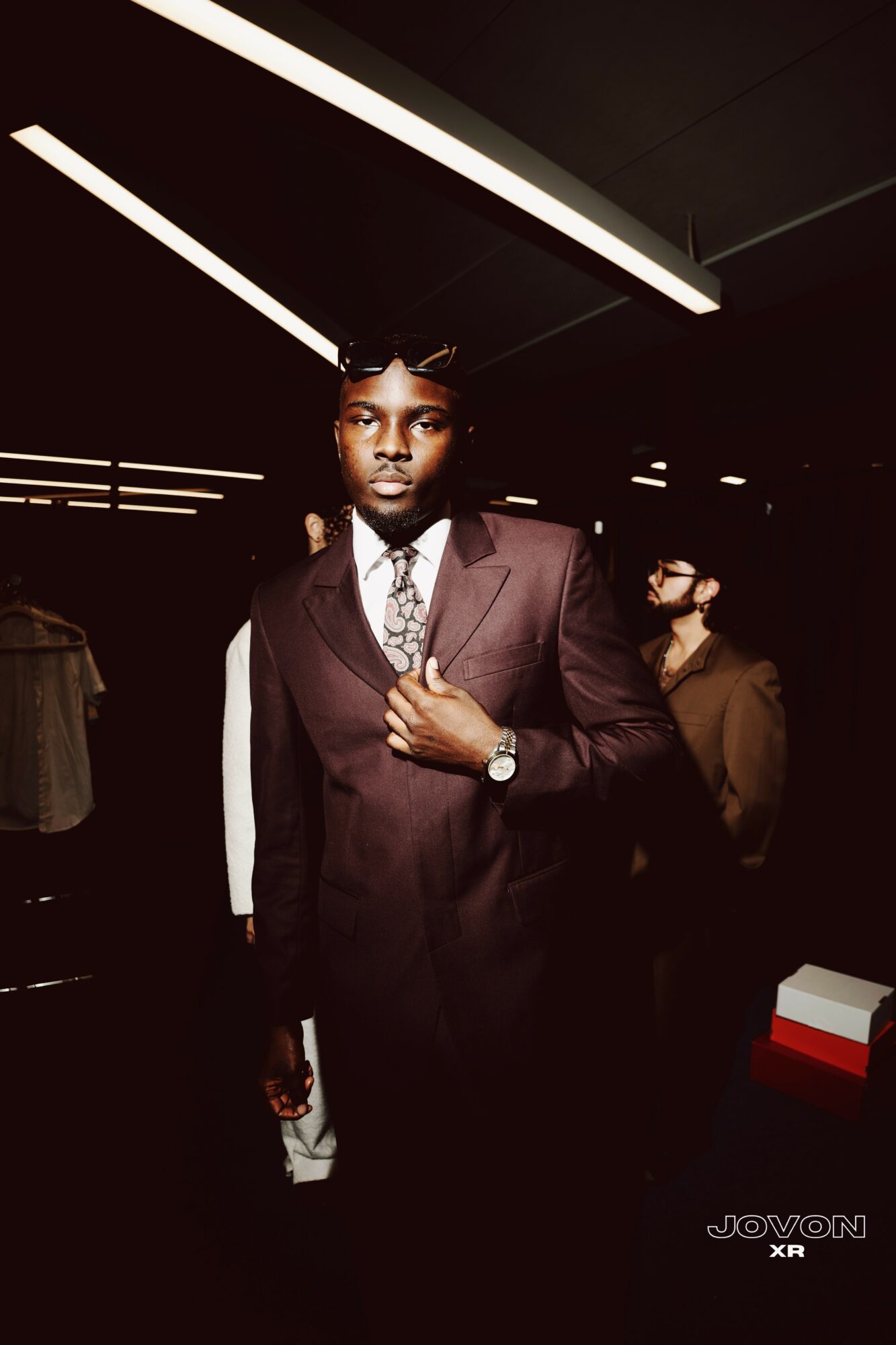 Young man in a dark suit and tie standing in a dimly lit indoor space with other people around.