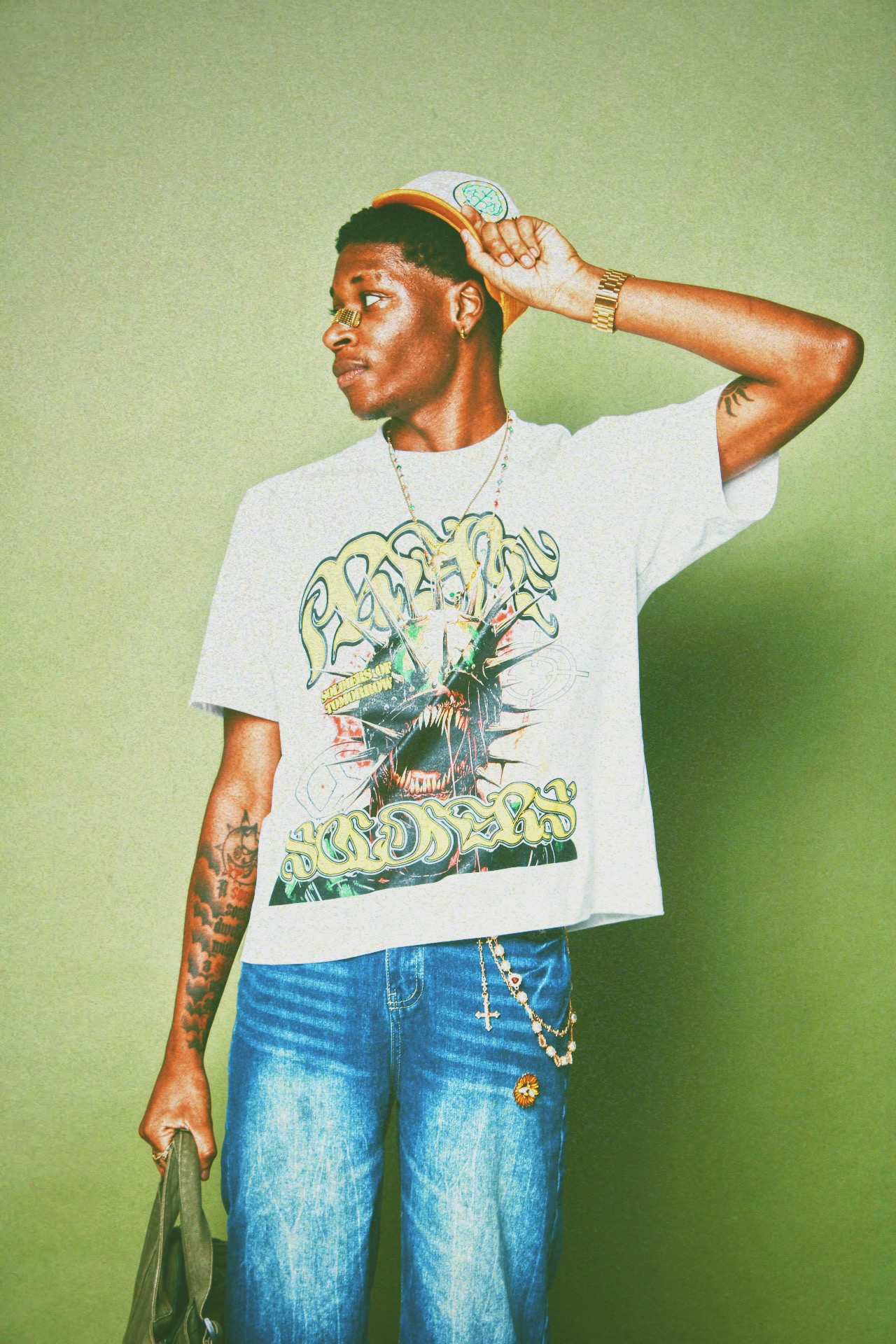 Young man with a cap, wearing a graphic t-shirt and jeans, holding a bag, standing against a green background.