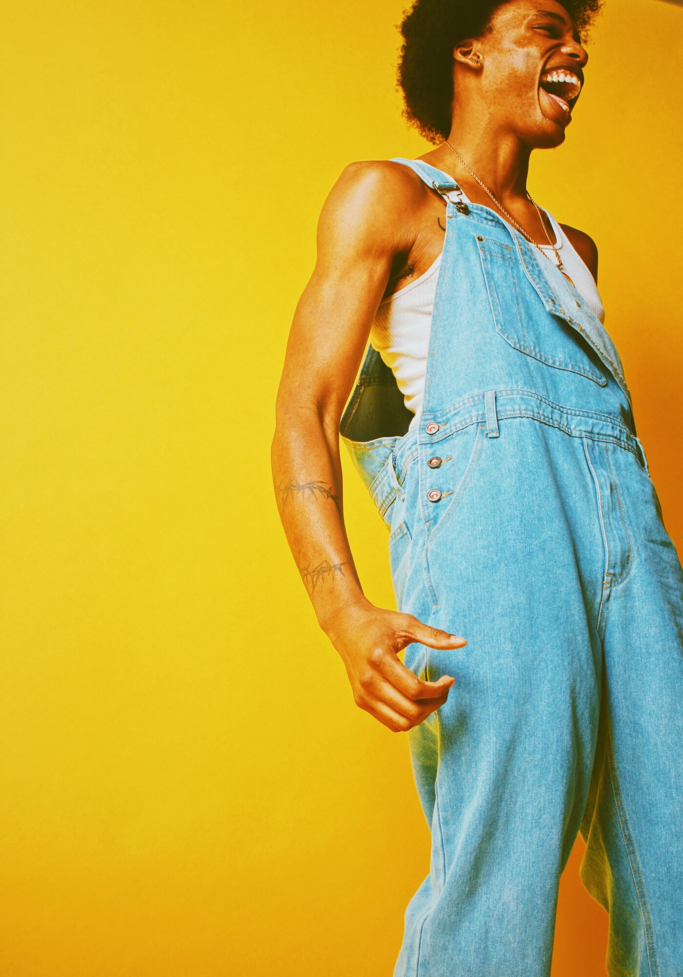Person laughing, wearing denim overalls and a white shirt, standing against a yellow background.