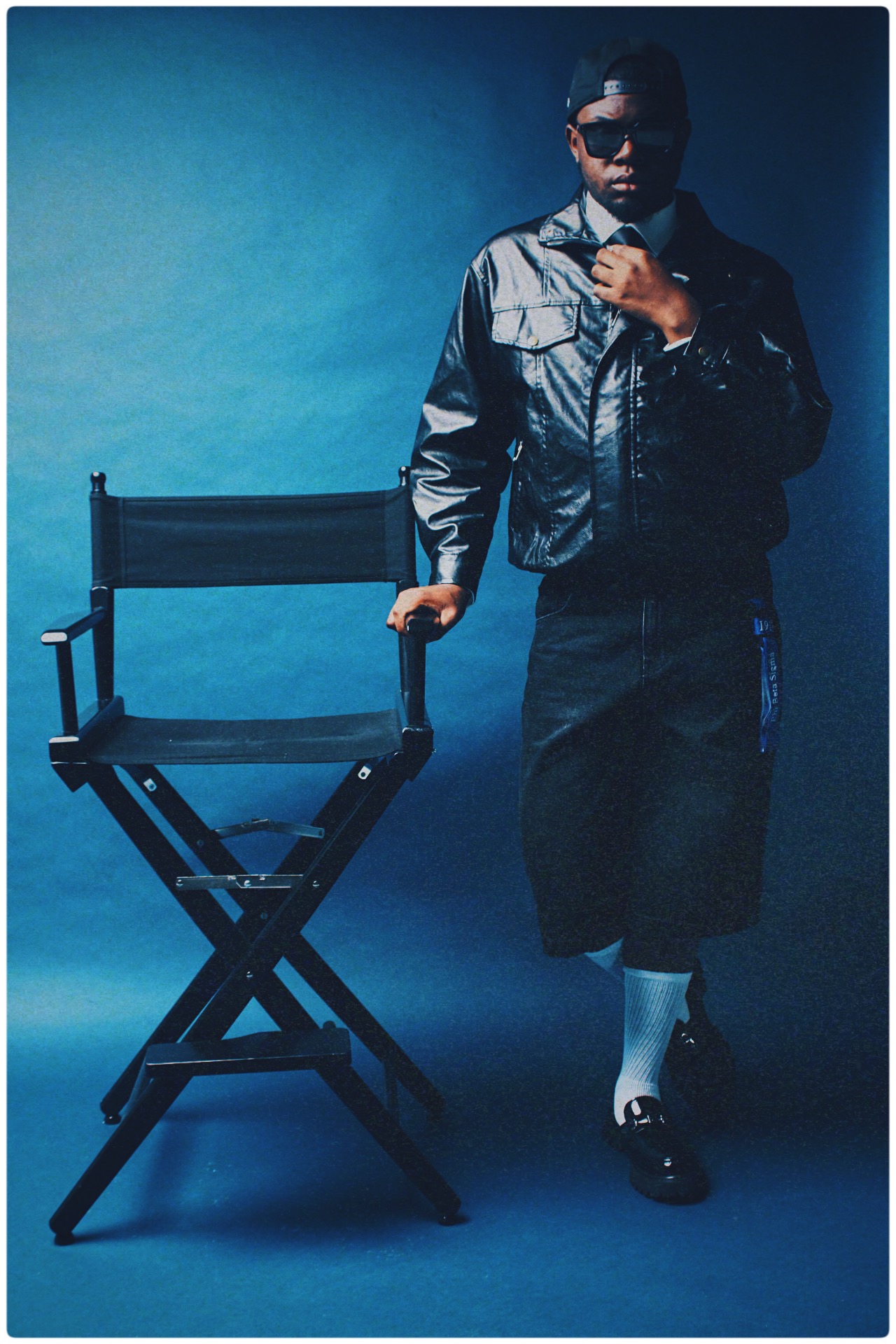 Person standing next to a director's chair, wearing sunglasses, a jacket, and a skirt, against a blue background.