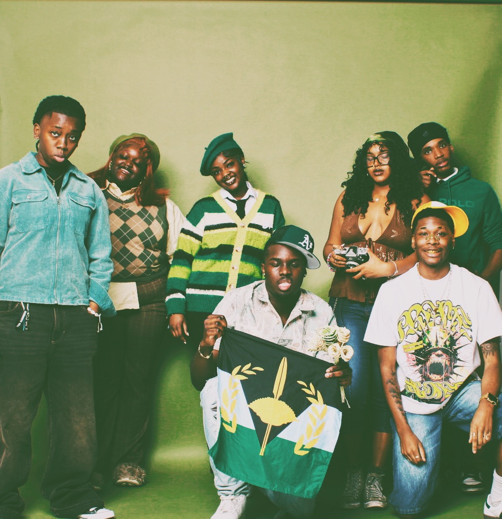 Group of eight diverse people standing against a green background, some holding a flag, smiling and looking at the camera.