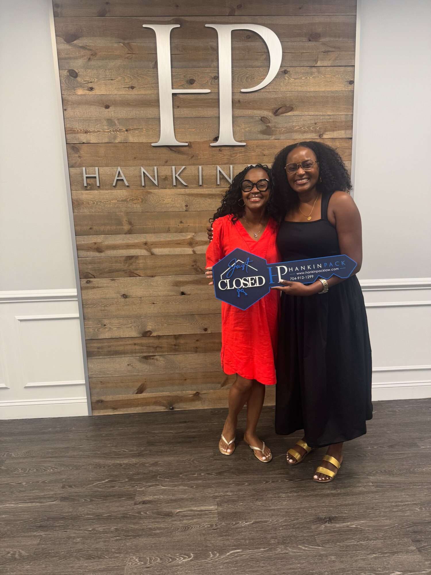 Two women stand in front of a wooden wall with large initials 'HP' and the word 'HANKINS'. One woman wears a red dress, the other a black dress, and they hold a large blue key-shaped sign.