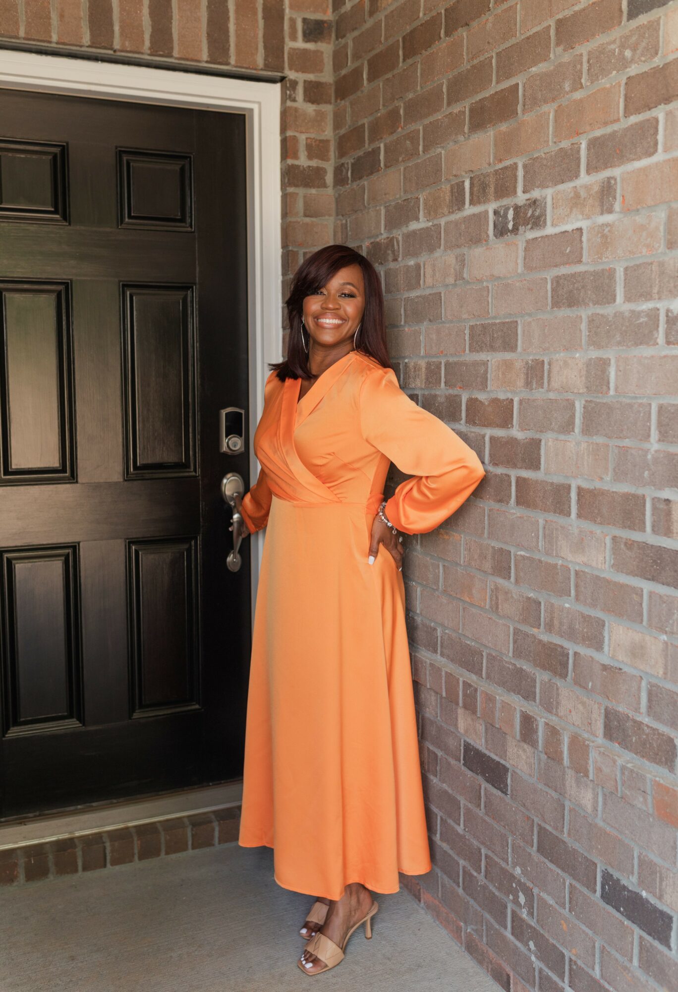 Woman in orange dress standing next to brick wall and black door, smiling, with hands on hips.