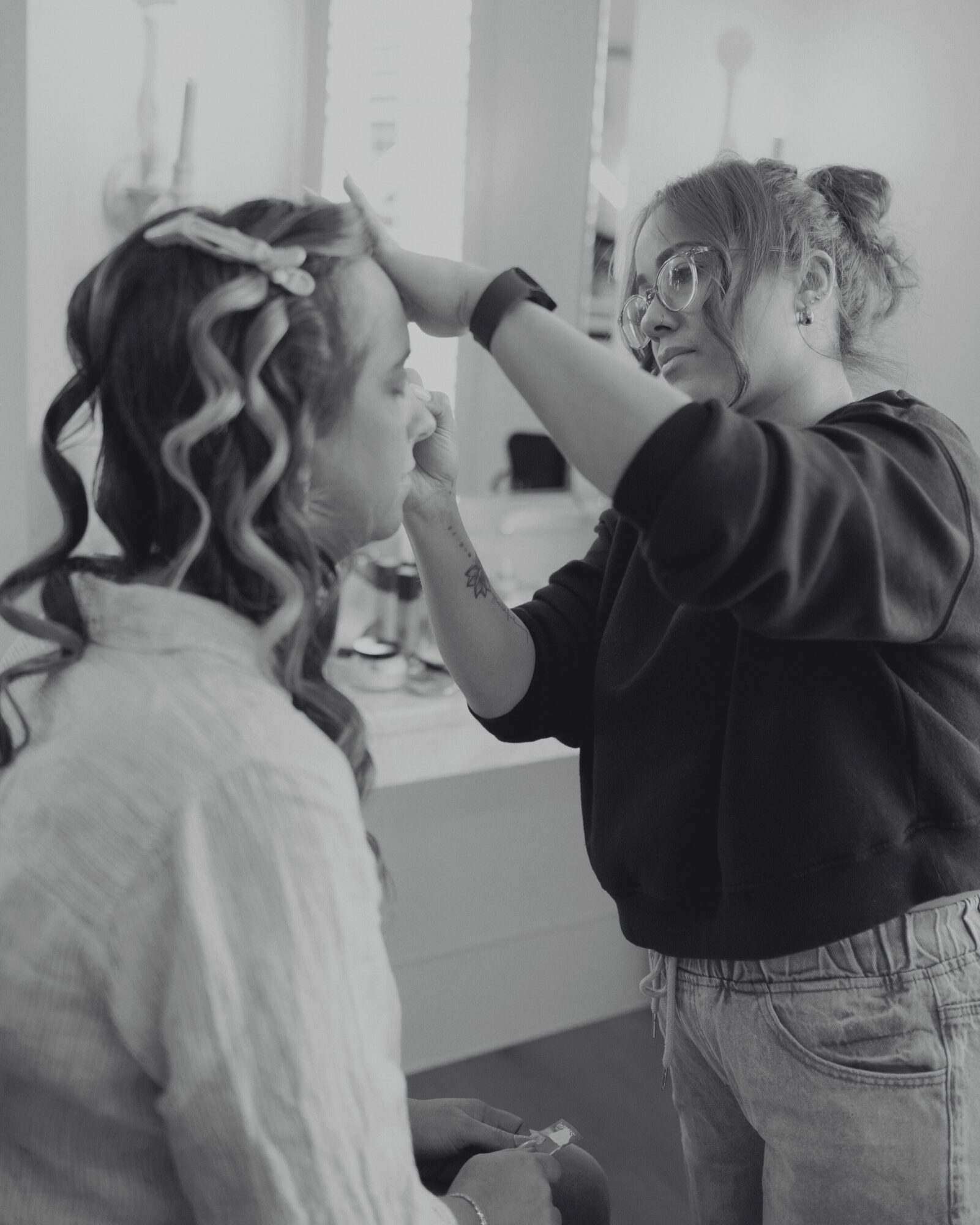 Makeup artist applying makeup to a woman with curled hair, in a room with a mirror and bright lighting.