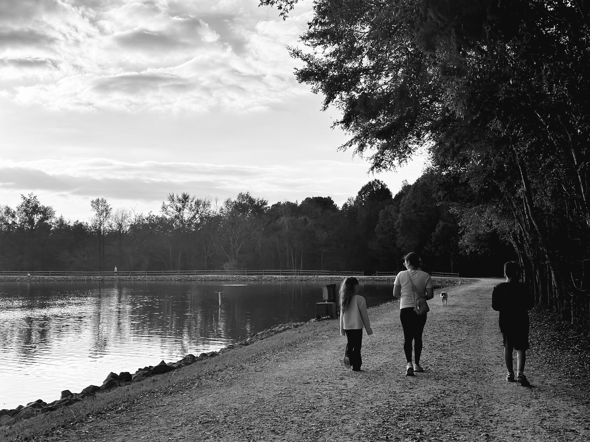 Three people walk along a lakeside path with trees on the right and water on the left, under a partly cloudy sky.