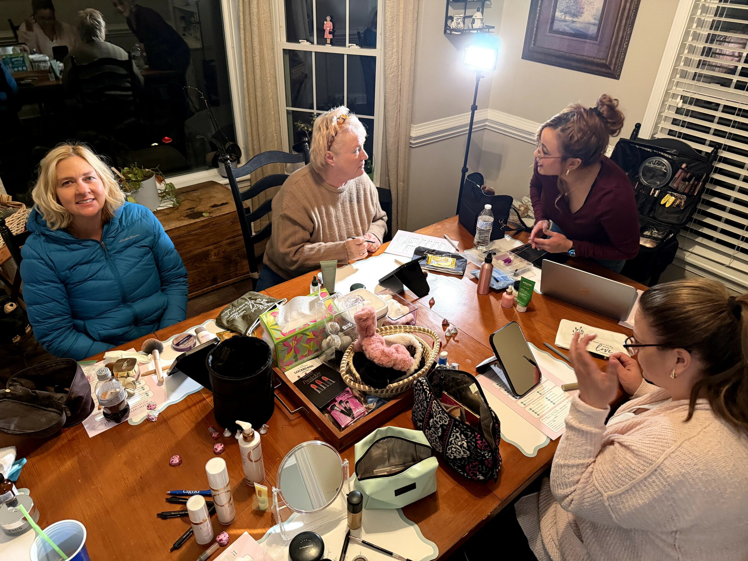 Four women sitting around a dining table engaged in conversation, with various items and a lamp in a cozy room.