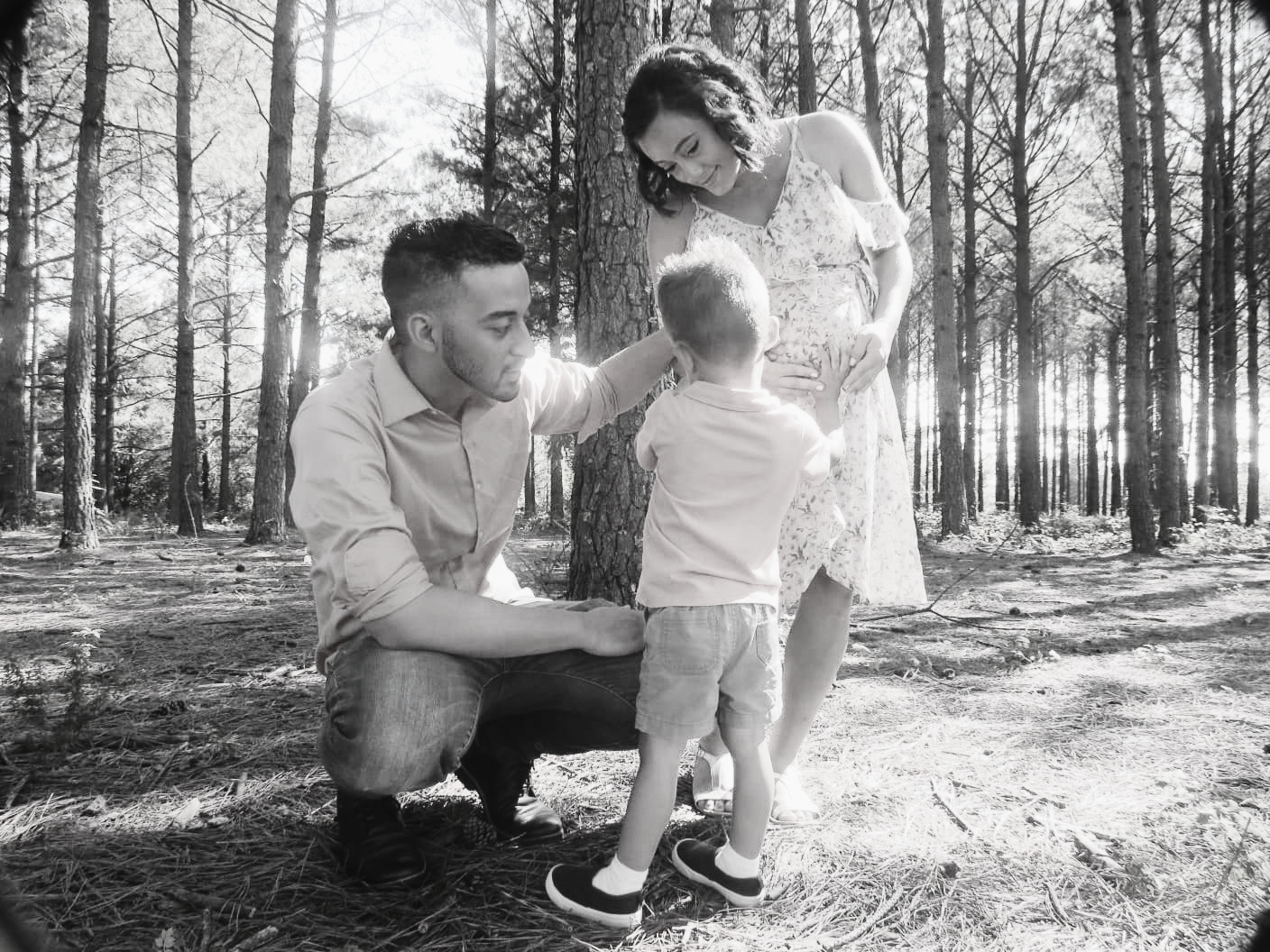 Family of three outdoors in a forest, with a man kneeling, a woman standing, and a child between them, trees in background.
