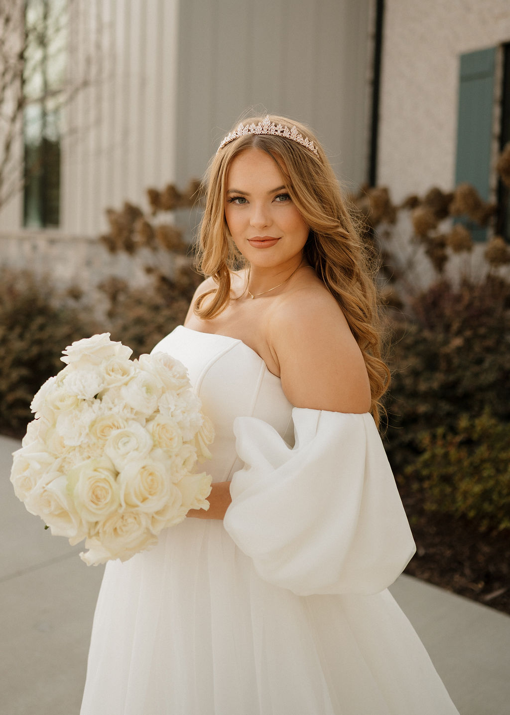 Young woman in white dress holding a large bouquet of white flowers, outdoors with bushes and buildings in background.