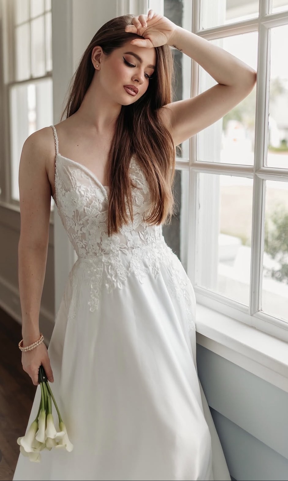 Young woman in a white dress standing by a window, holding flowers, with her hand on her forehead.