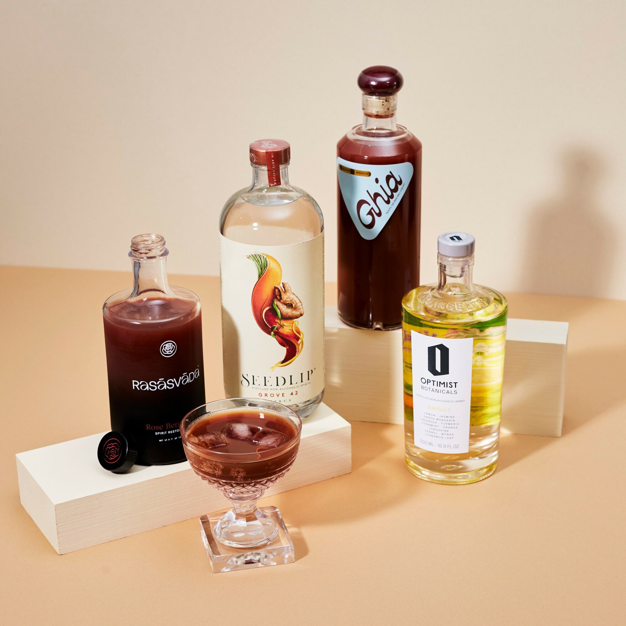 Four bottles of different beverages and a glass with a dark drink on a white platform, against a beige background.