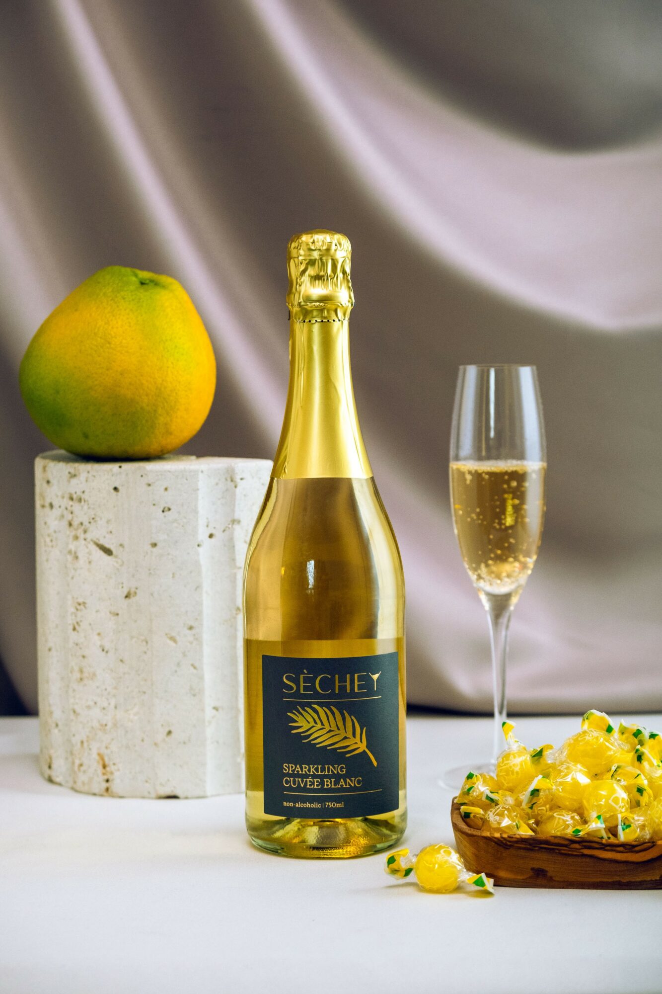 A bottle of sparkling wine, a glass of champagne, a green apple on a marble pedestal, and a dish with yellow flowers.