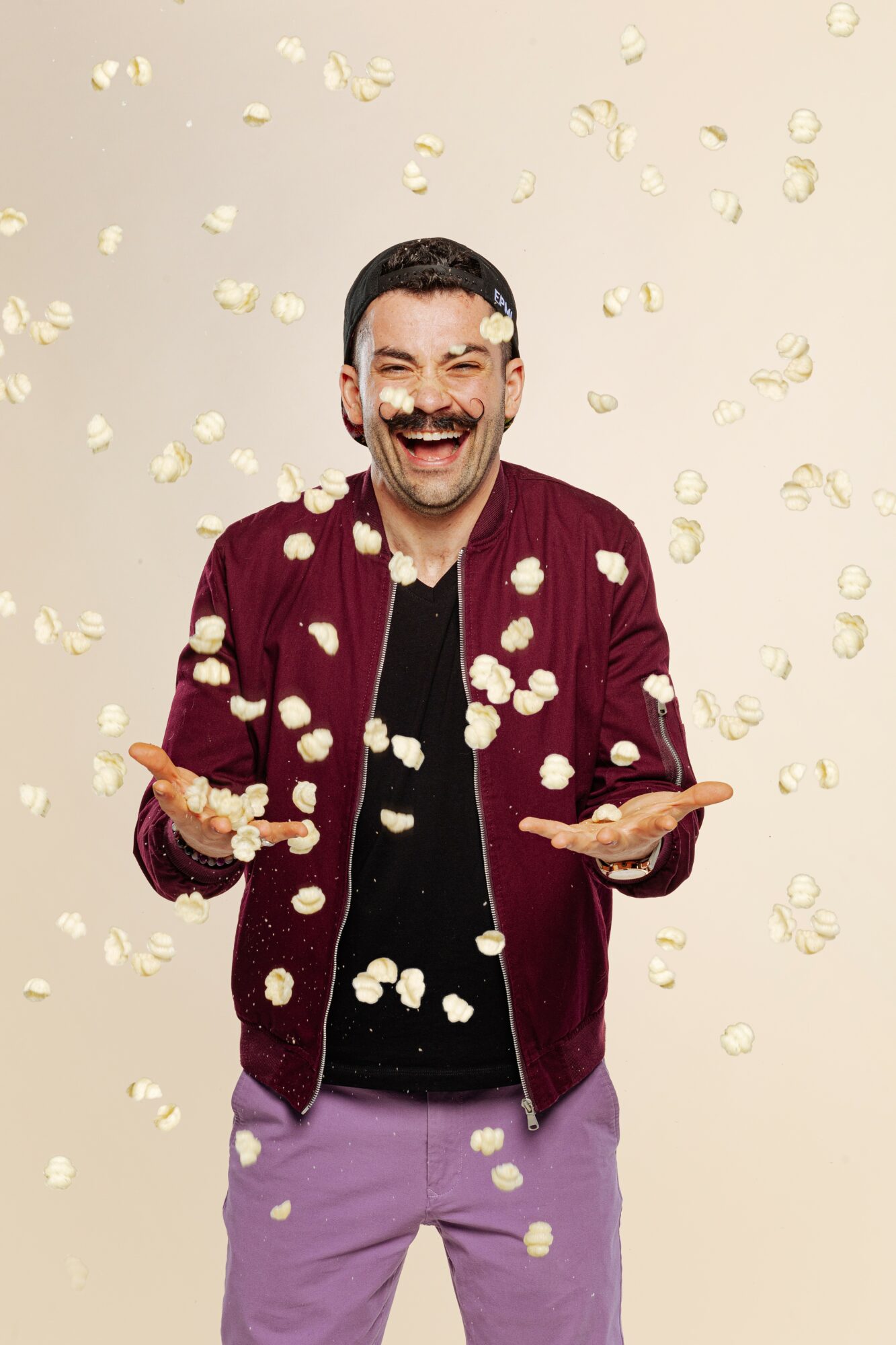 Man smiling with arms outstretched, surrounded by falling popcorn, wearing a maroon jacket and purple pants.