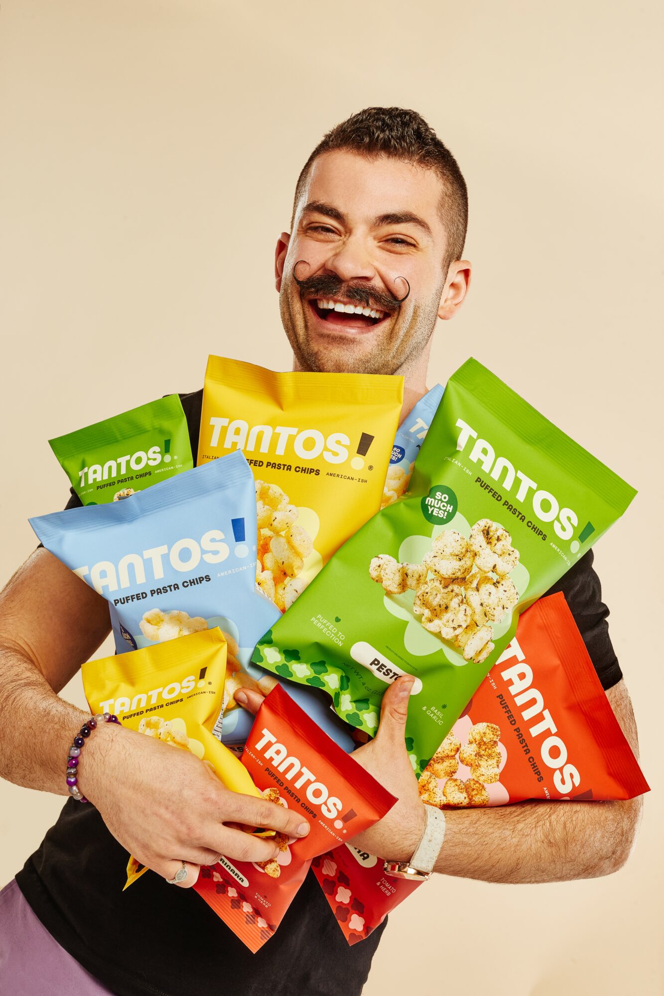 Man smiling and holding multiple bags of TANTOS popcorn in various colors against a plain background.