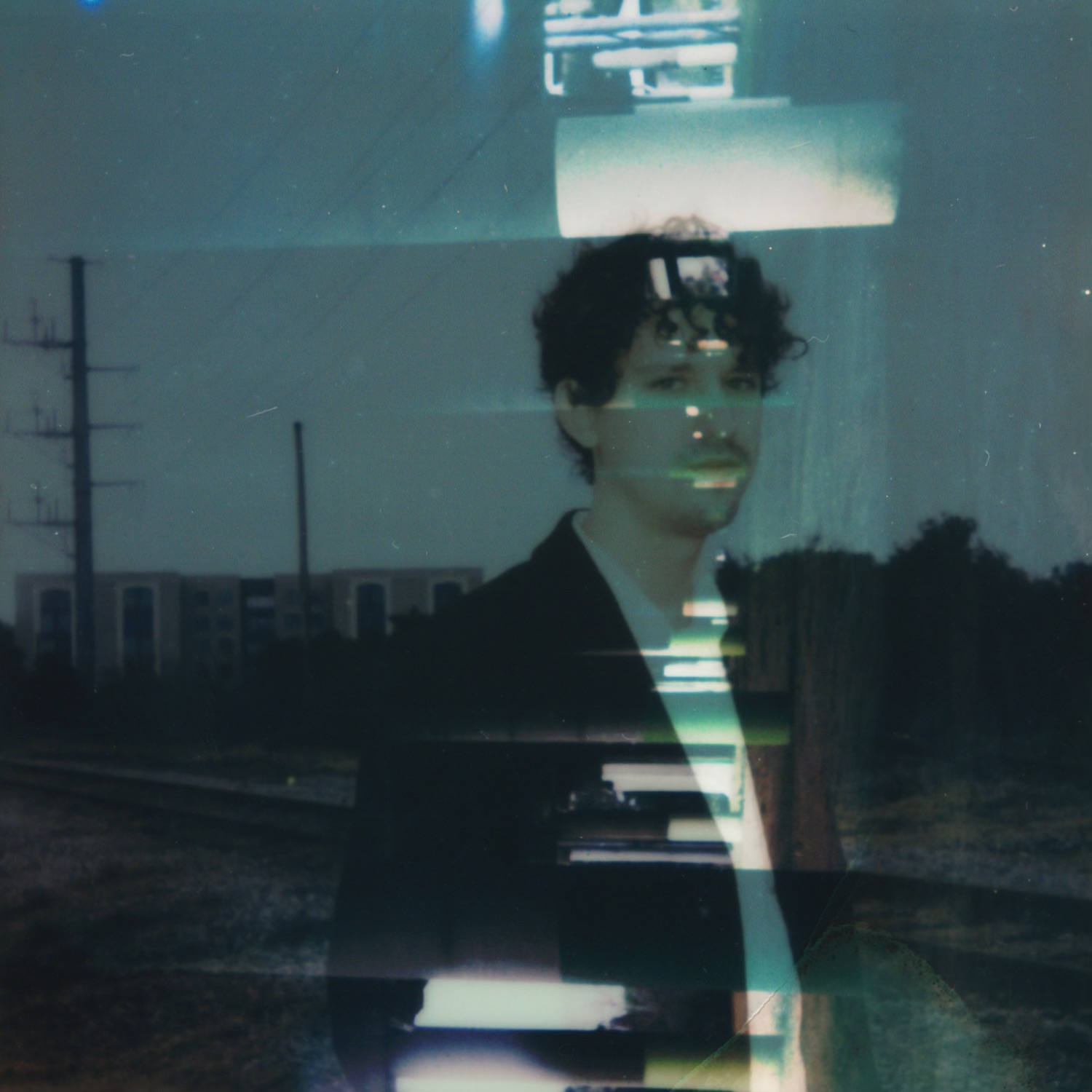 A person with curly hair stands outdoors near train tracks, with a cityscape and power lines in the background.