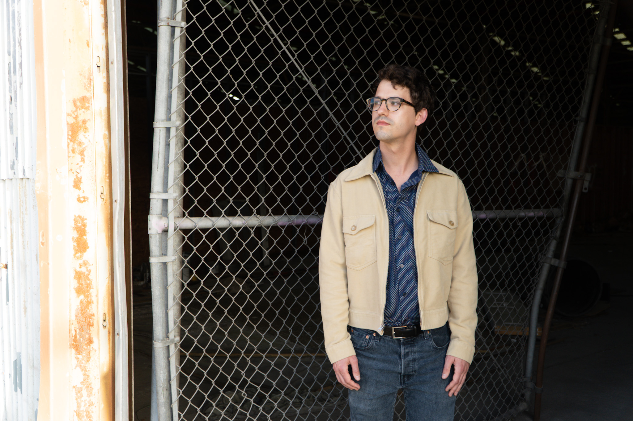 Man standing outdoors near a chain-link fence, wearing glasses, a beige jacket, and blue shirt, looking to the side.