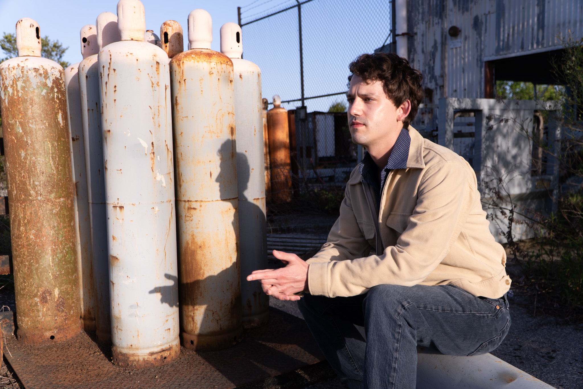 Young man crouching outdoors near rusted gas cylinders, industrial background, sunlight, casual clothing, focused expression.