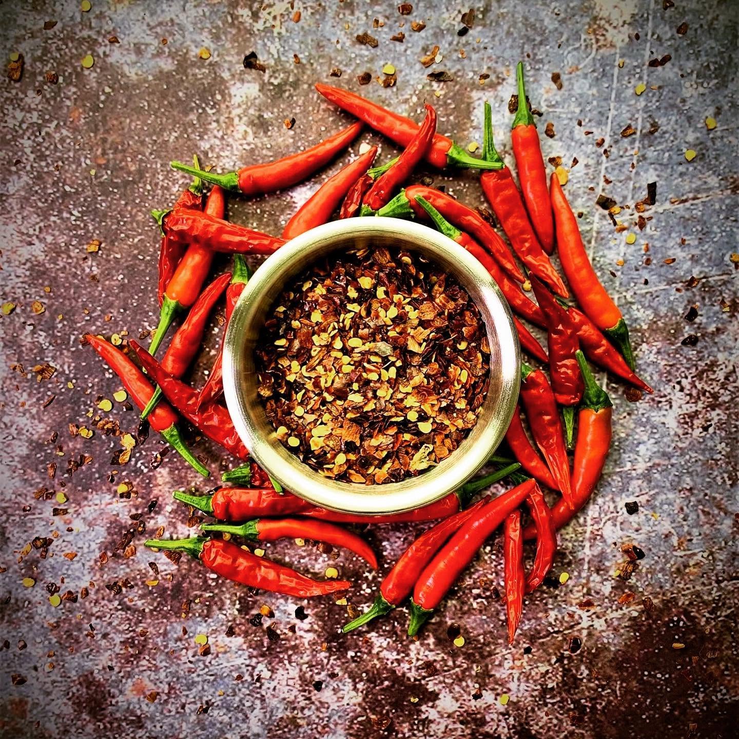 Red chili peppers surrounding a bowl of crushed chili flakes on a textured surface.