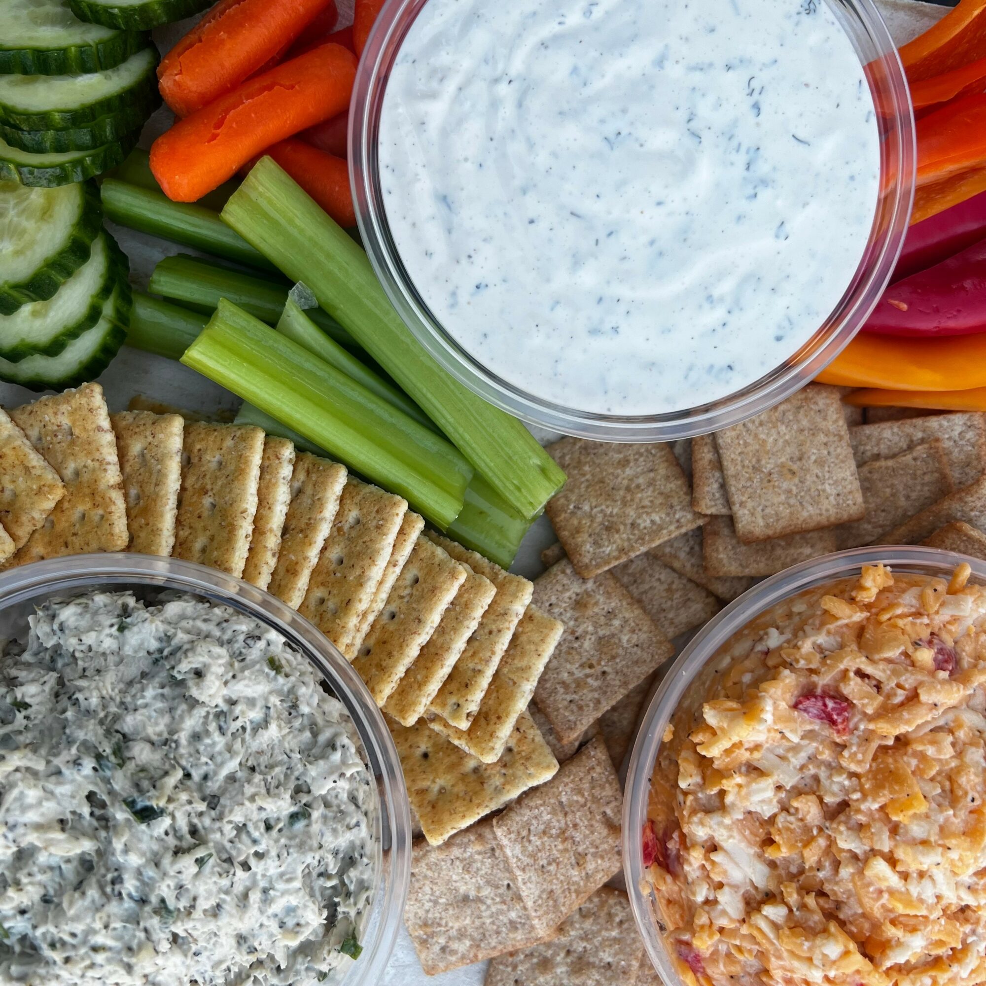 Vegetable platter with dip, crackers, and spread, including cucumber, carrots, celery, and colorful bell peppers.